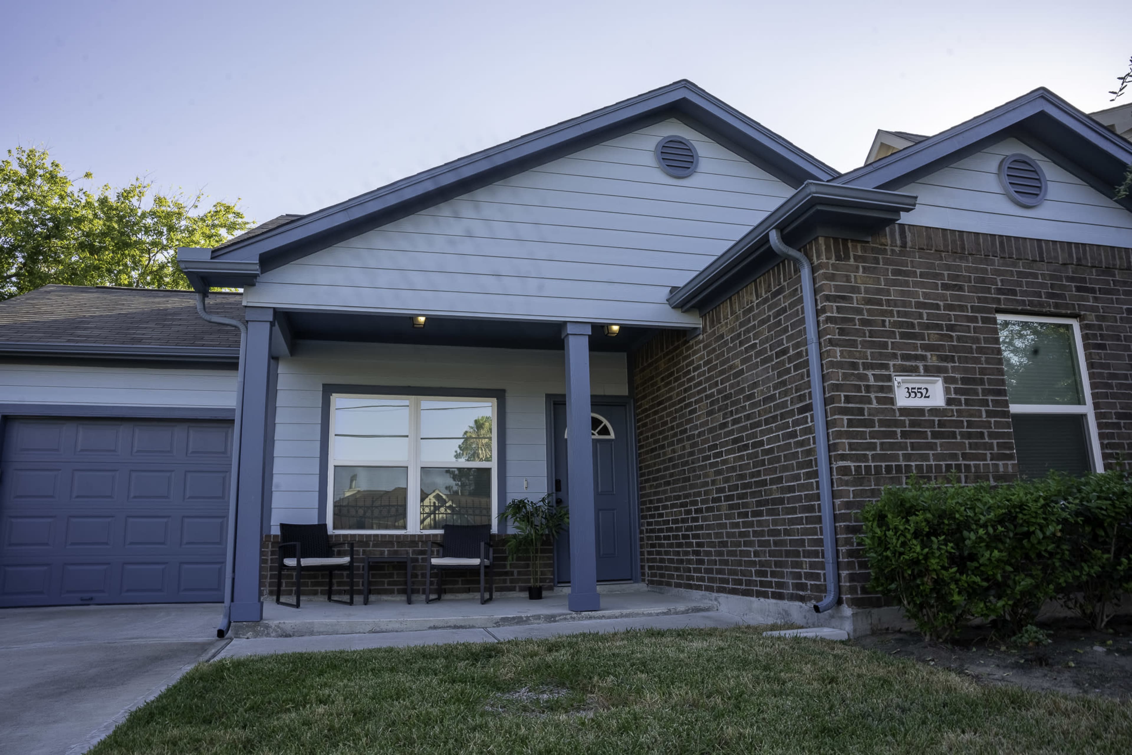 A single-story house with a brick facade and light blue siding, featuring a small porch with two chairs and a garage on the left side.