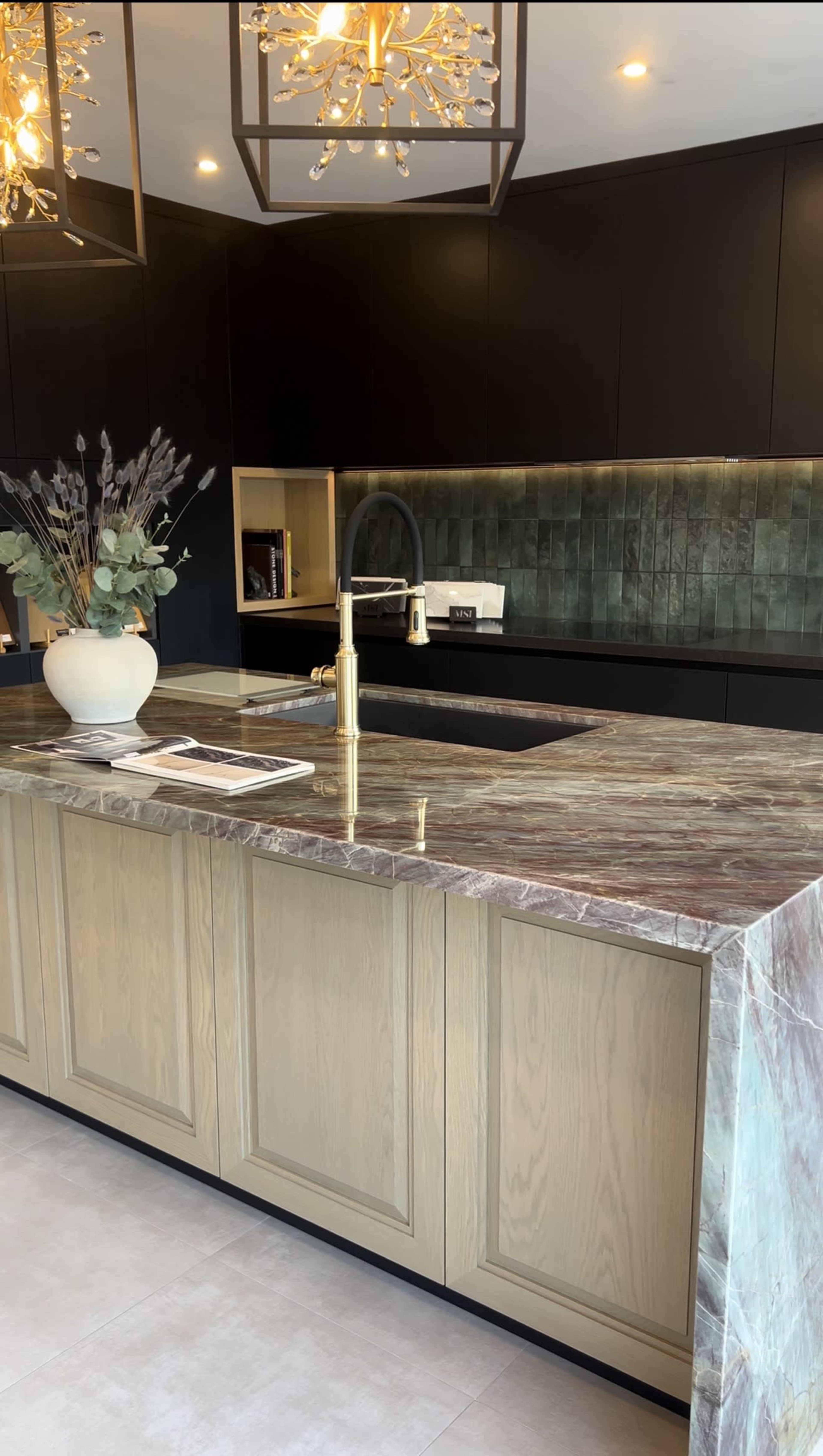 The image shows a modern kitchen with a large marble countertop, dark cabinetry, a gold faucet, and decorative lighting fixtures.