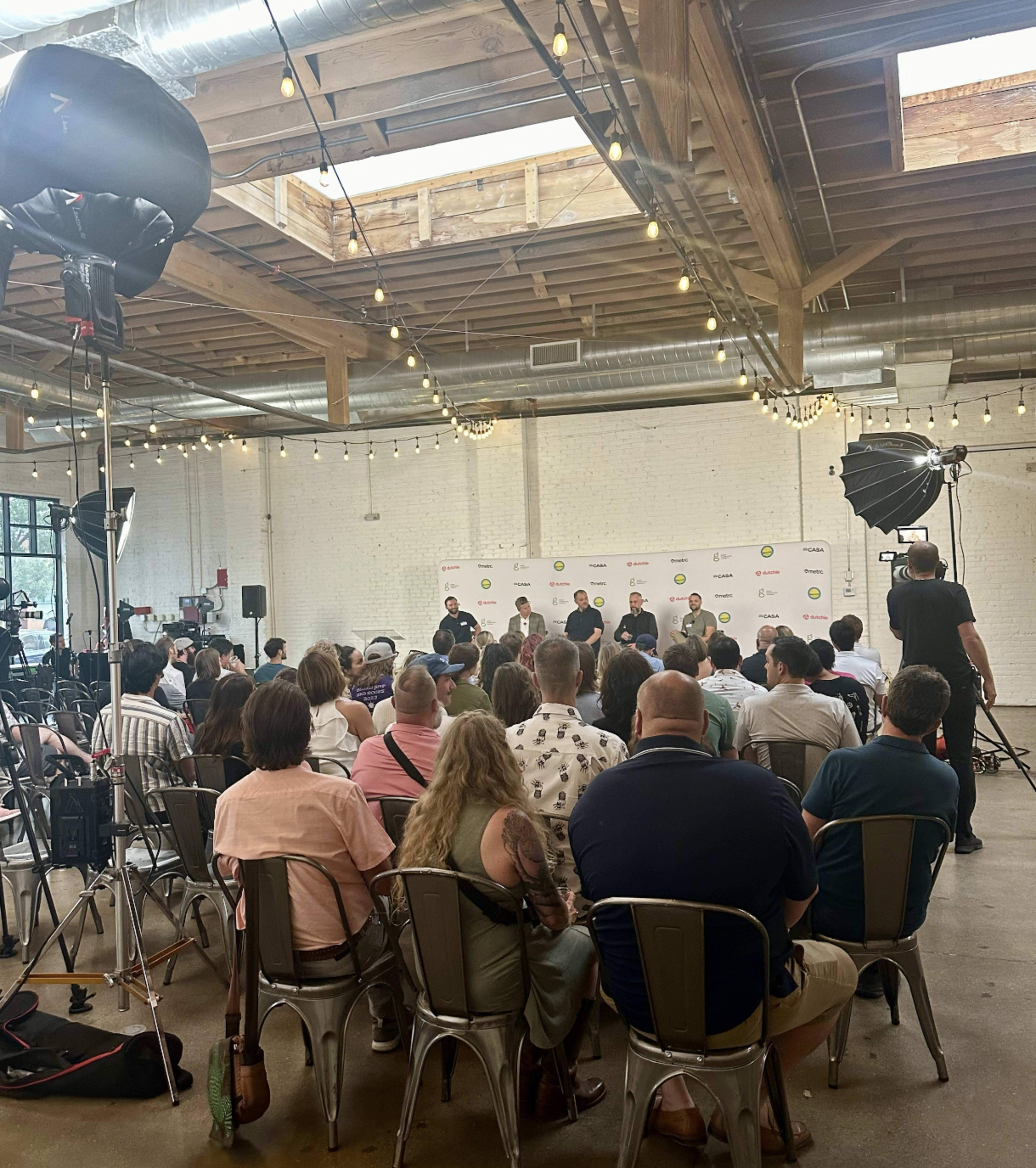 A panel discussion takes place in a well-lit venue with a large audience seated on metal chairs, while several cameras and lights are set up for recording.