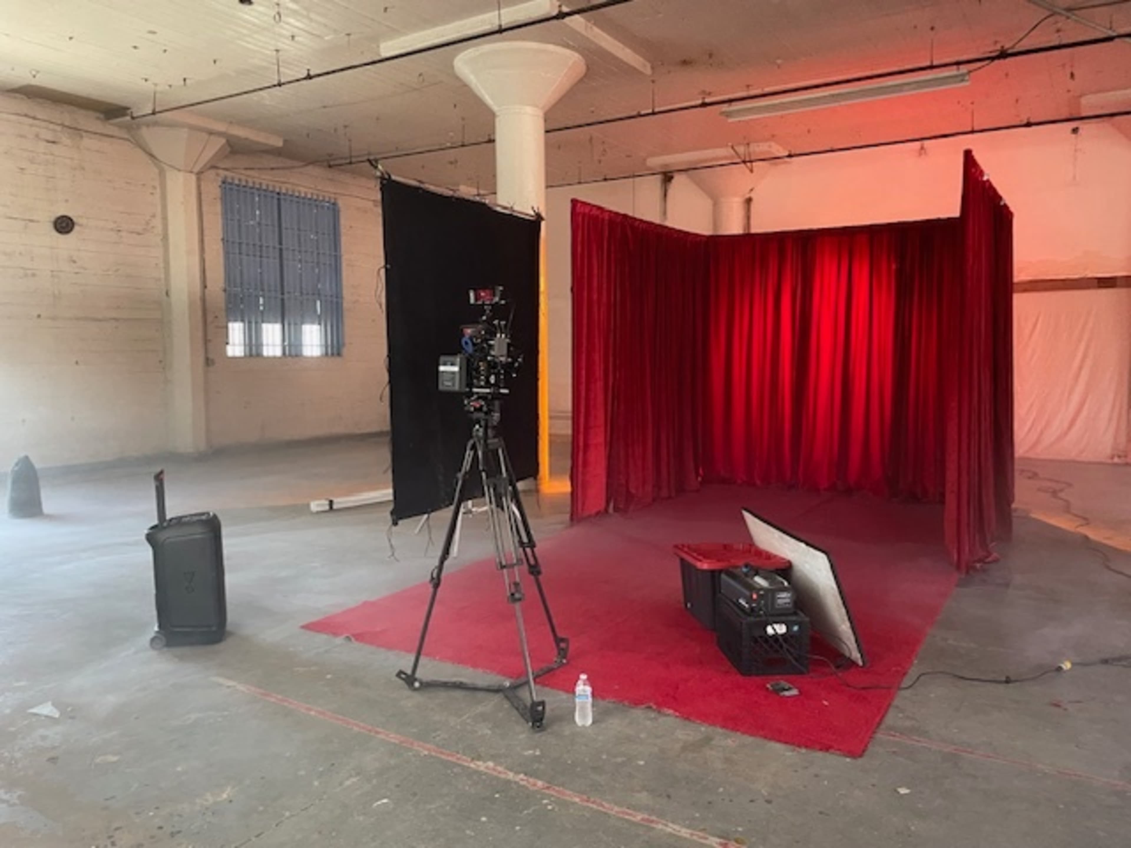 A camera is set up on a tripod in front of a red velvet backdrop, with a piece of red carpet and some equipment scattered in a spacious, industrial room.