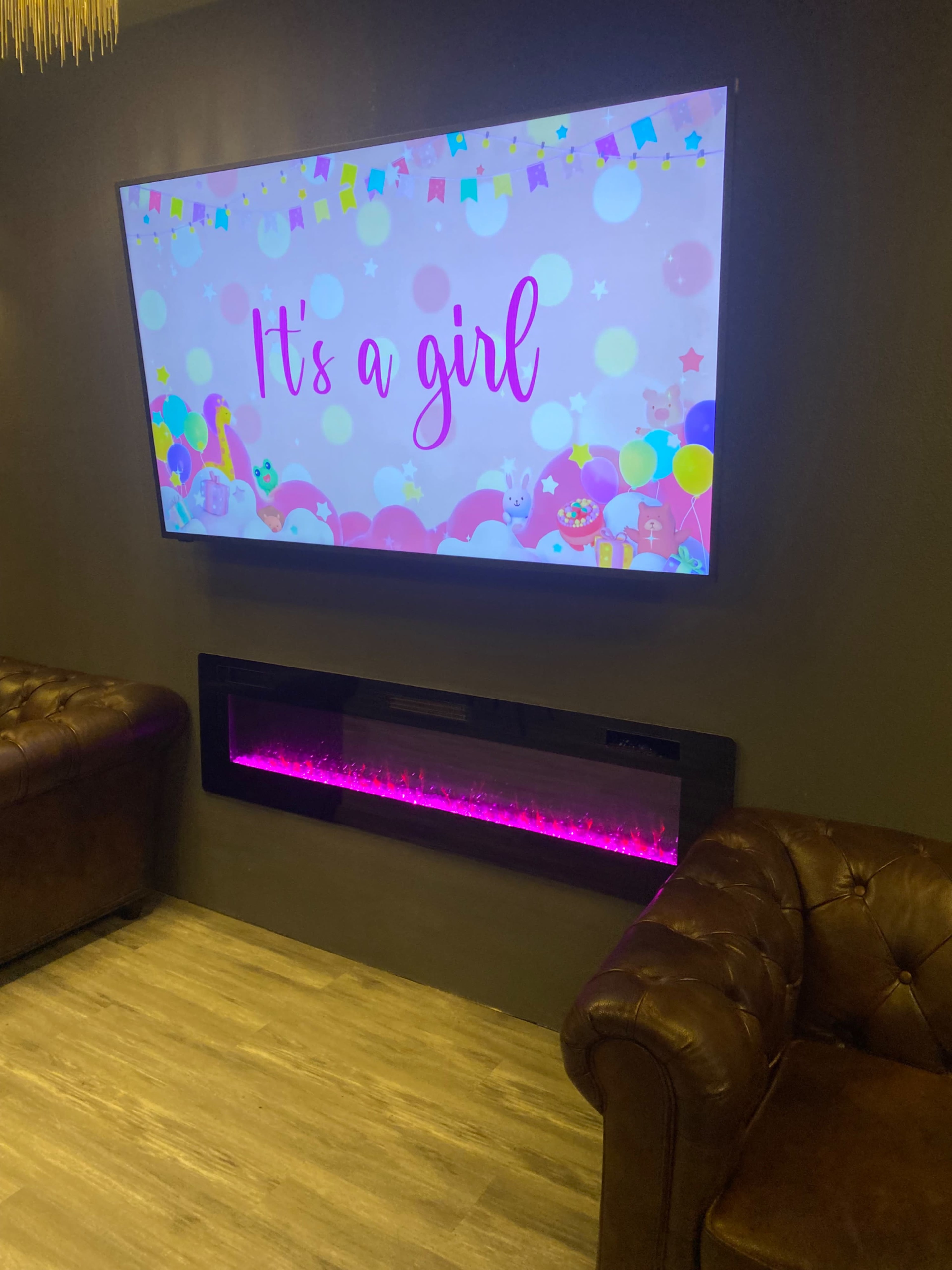 The image features a wall-mounted television displaying a colorful announcement that reads "It's a girl," above a modern electric fireplace with pink accent lighting, set in a room with two brown leather couches.
