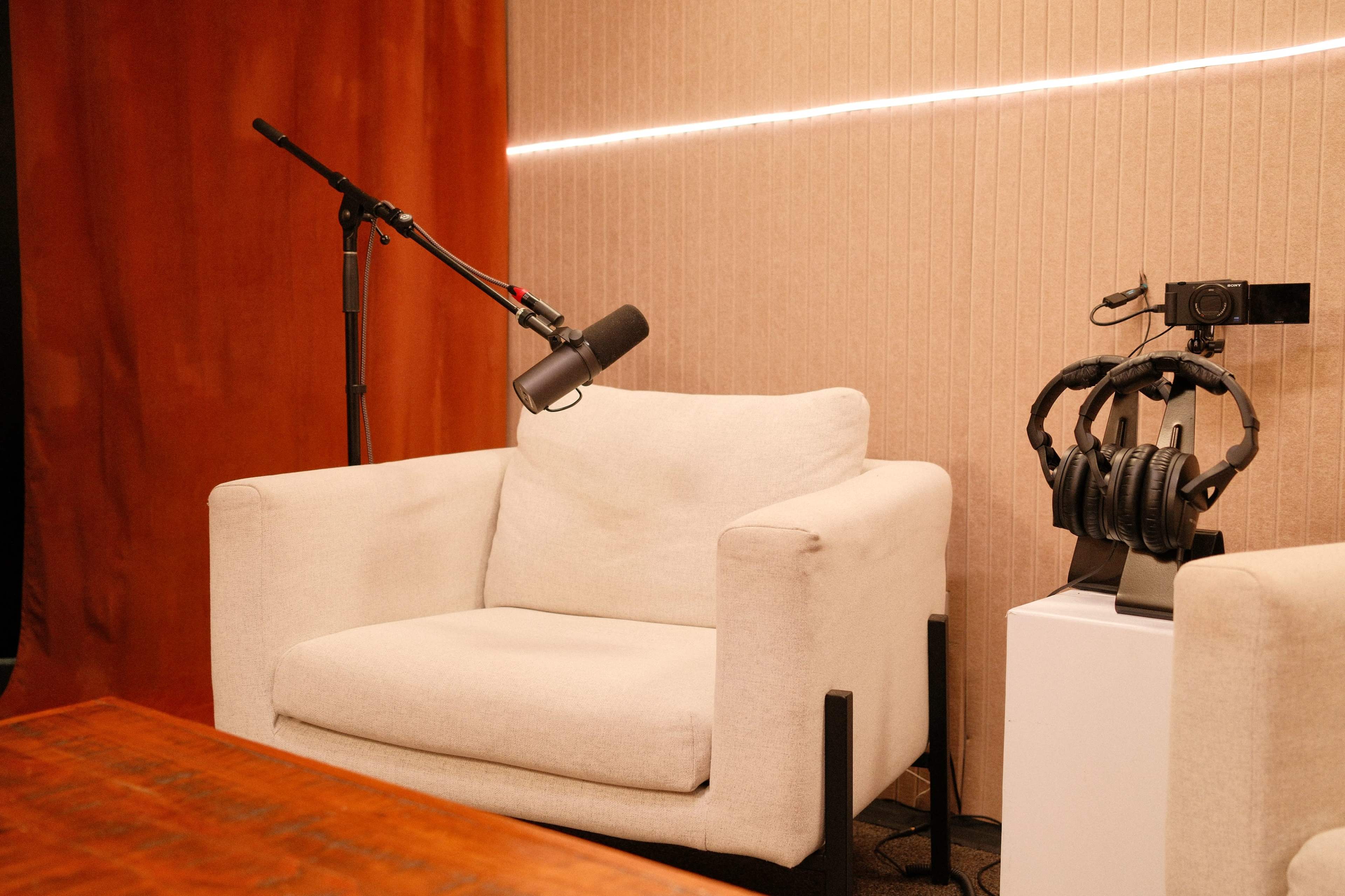 A microphone on a stand is positioned next to a white armchair, with headphones resting on a stand nearby against a backdrop of textured walls and ambient lighting.