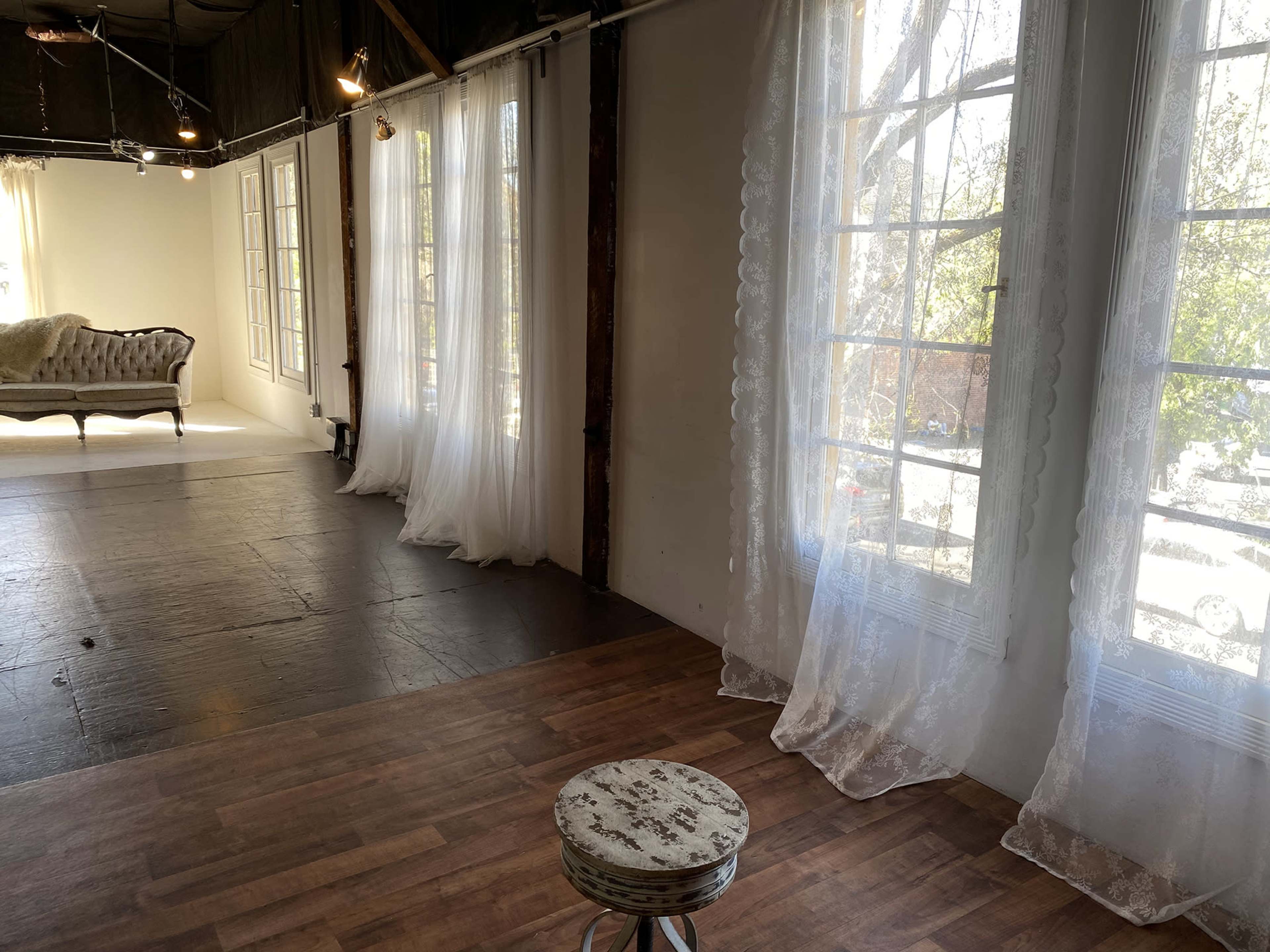 The image shows a bright, spacious room with large windows dressed in sheer curtains, a small round stool in the center, and a vintage sofa in the background.