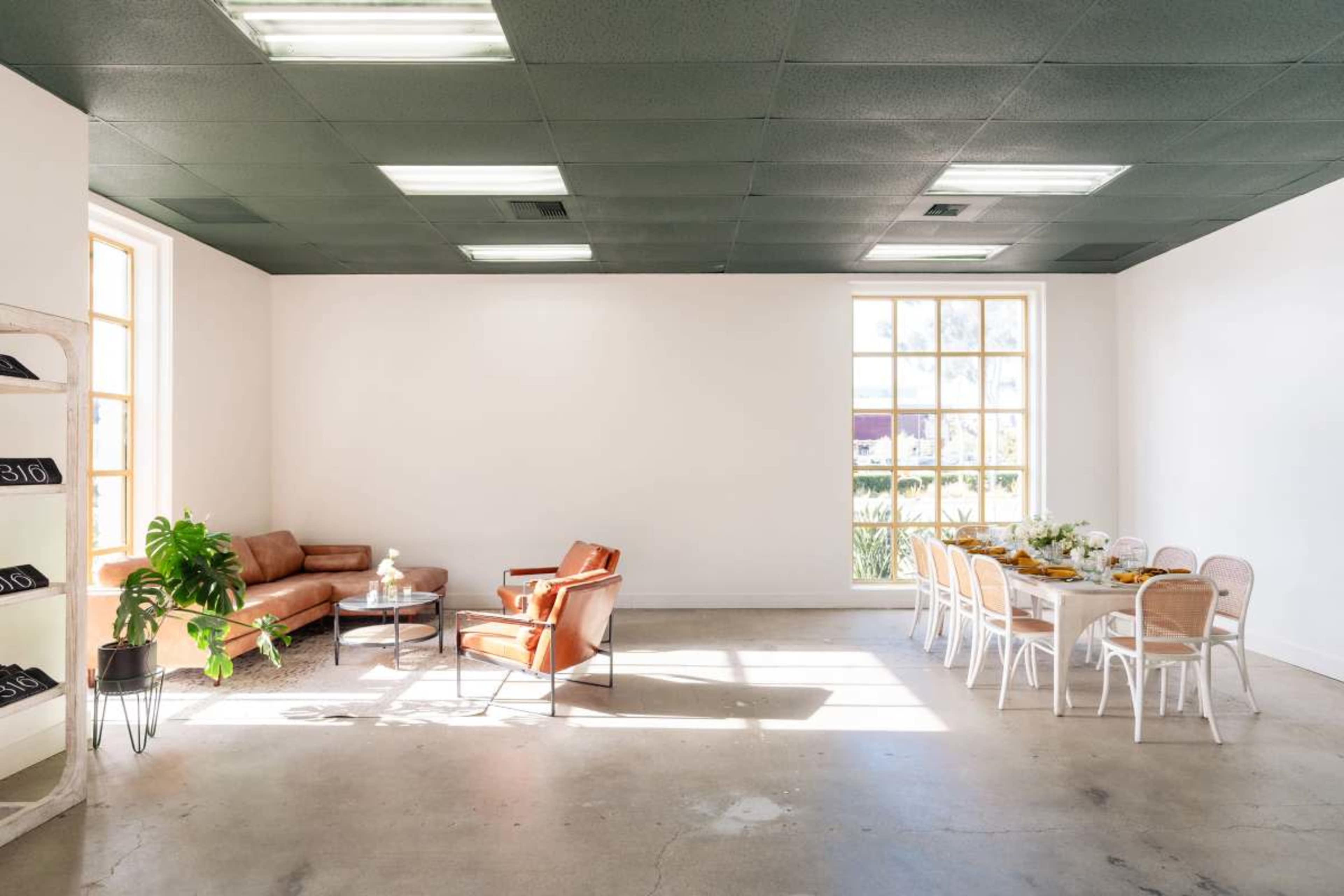 The image shows a bright, minimalist room featuring a brown leather sofa, an orange armchair, a small round table, and a dining table set for several people along with large windows letting in natural light.