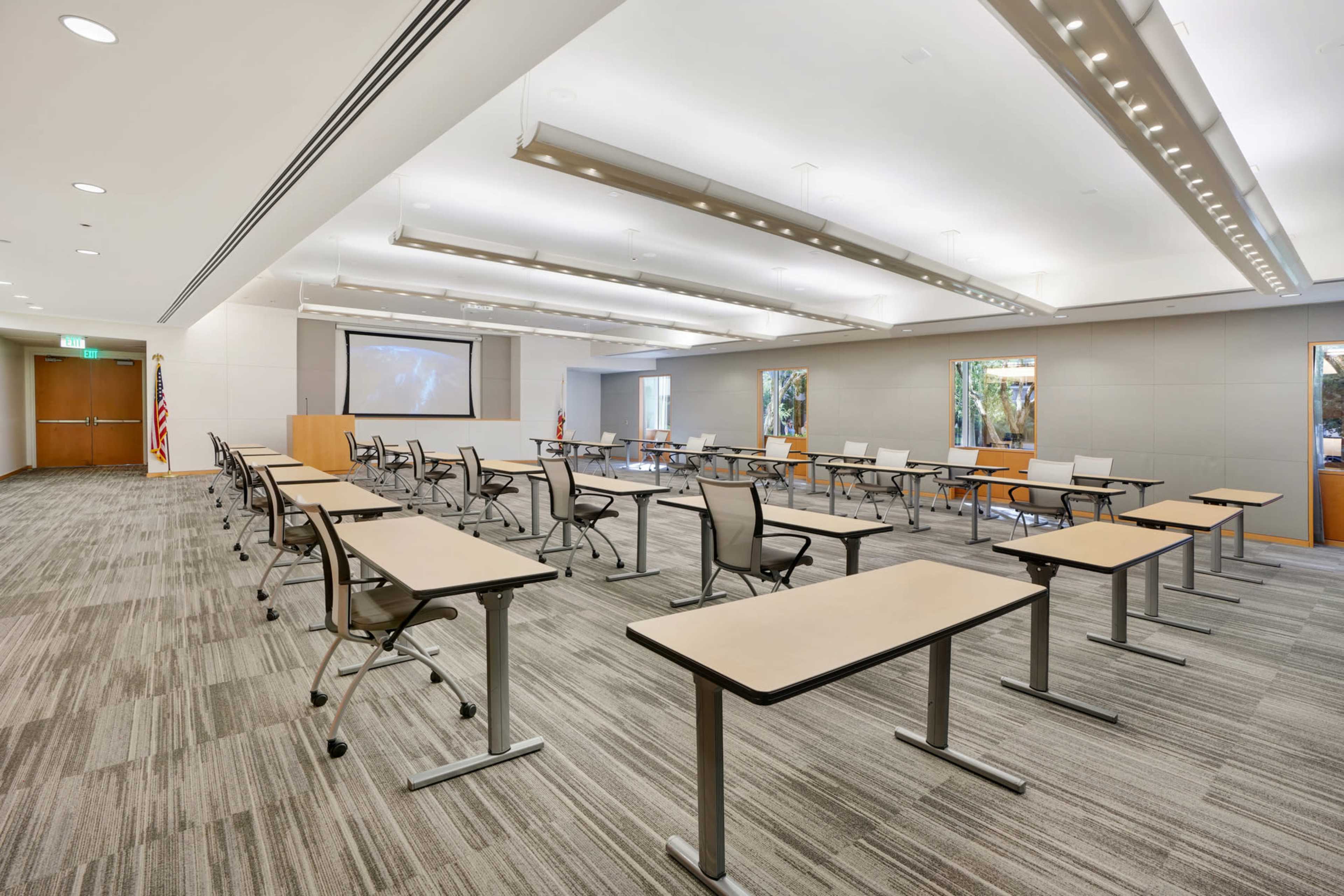 The image shows a spacious, modern classroom setup with multiple tables and chairs arranged in rows, along with a projector screen at the front.