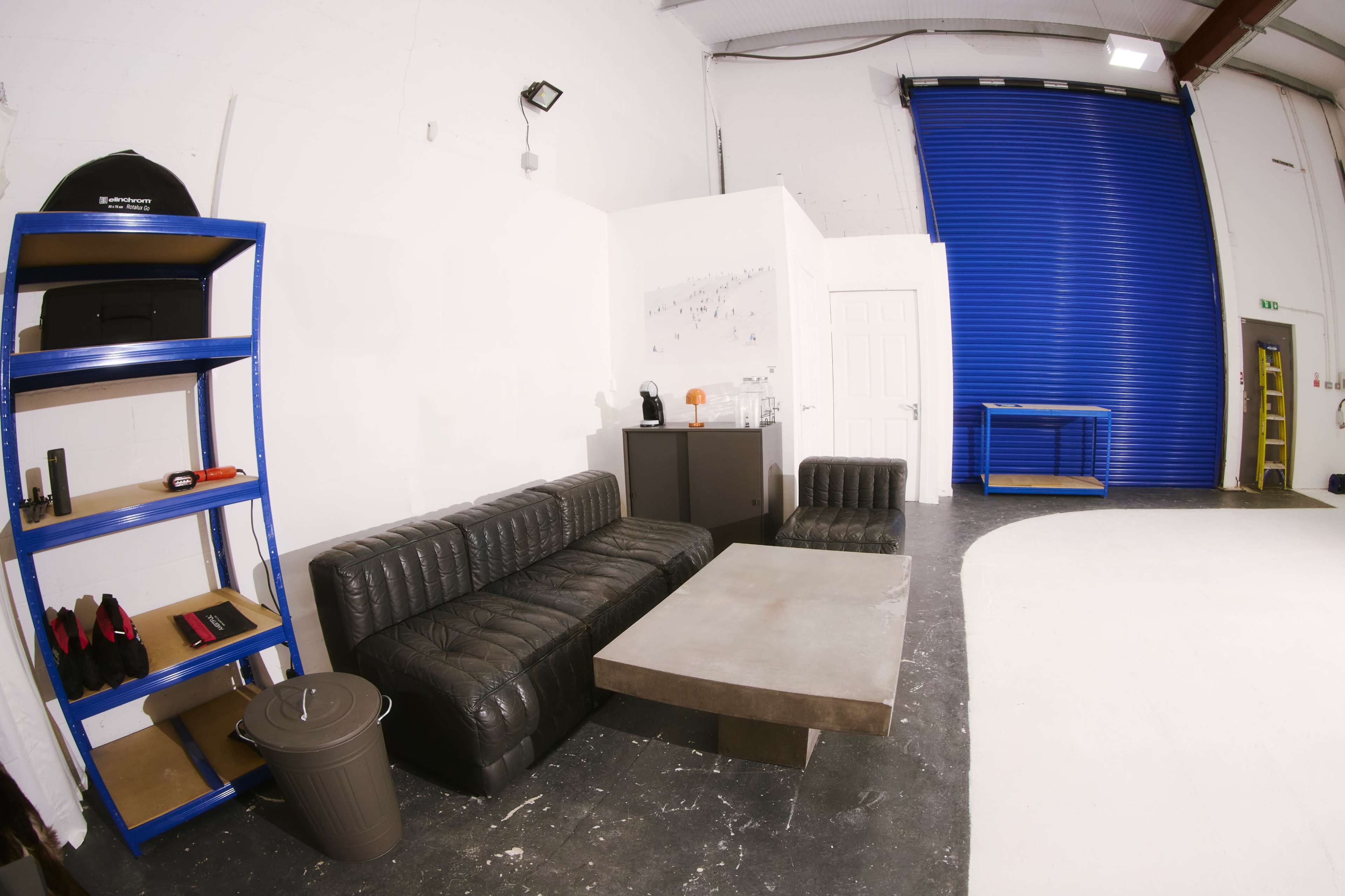 A spacious indoor area featuring a black leather couch, a small armchair, a metal shelving unit, a coffee table, and a door beside a large blue roll-up door.