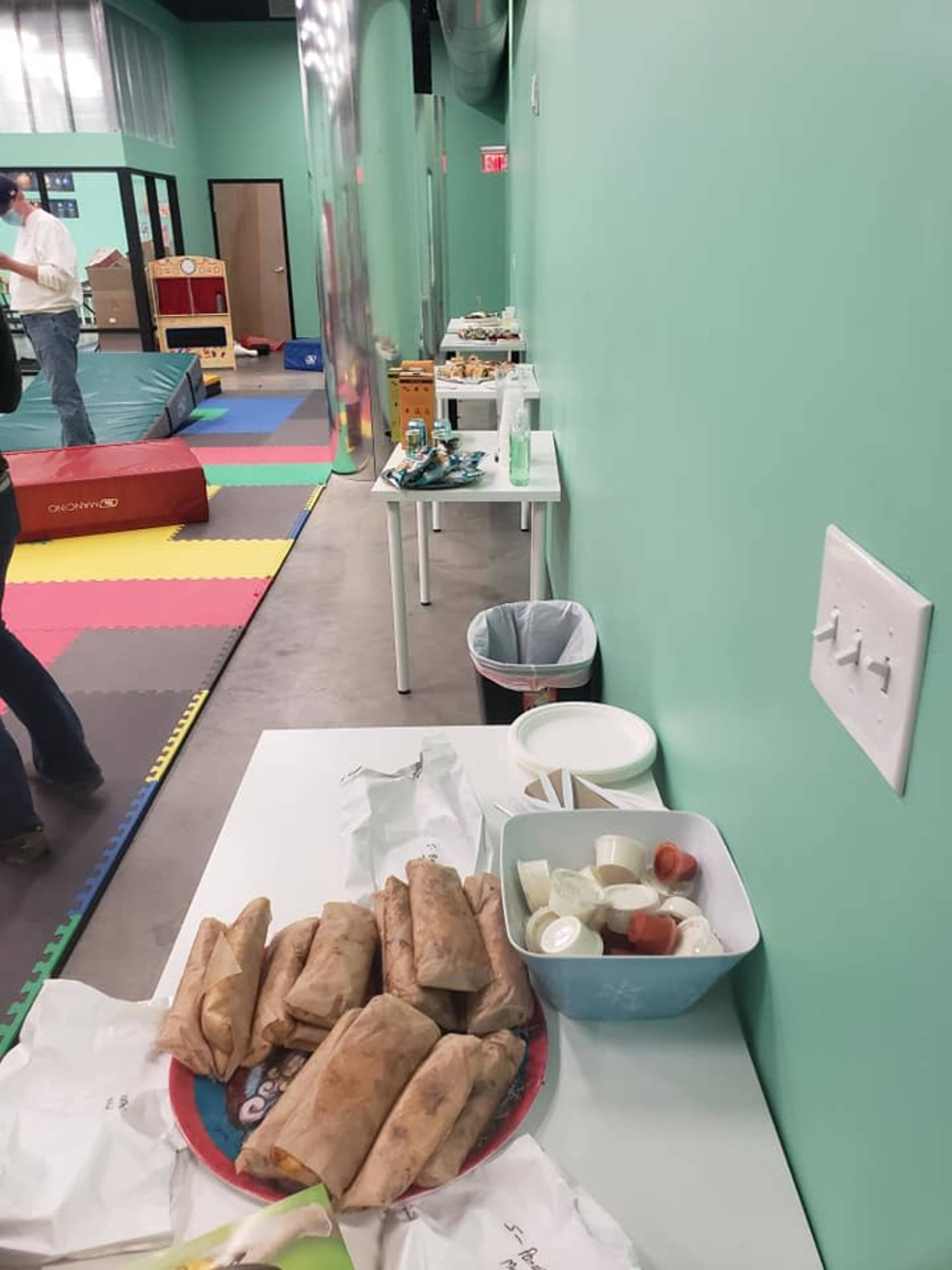 A green-walled room with a table displaying food items, including wrapped burritos and condiments, alongside a play area visible in the background.