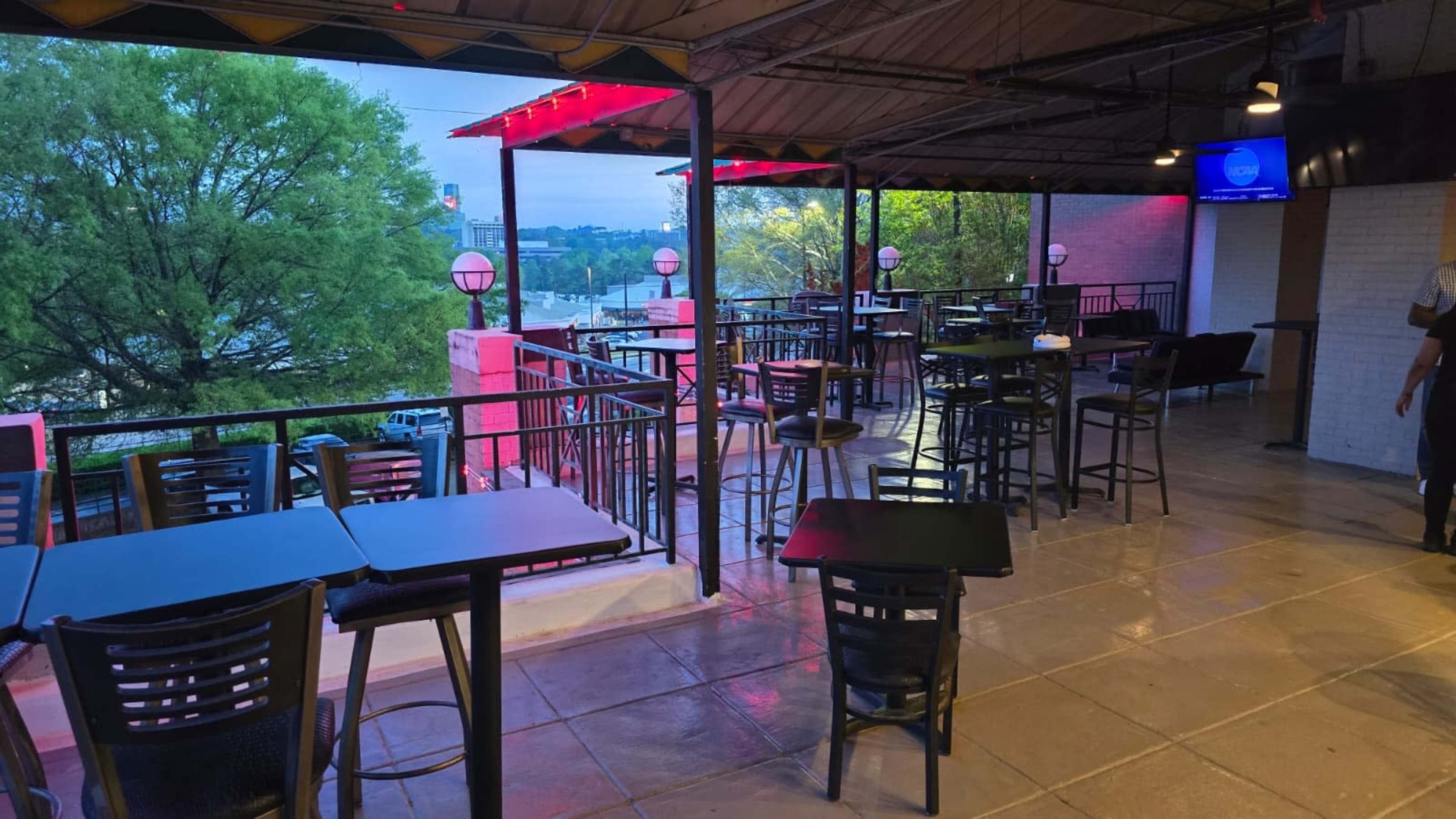 The image shows a restaurant or bar with an outdoor seating area equipped with black tables and chairs, overlooking a view of trees and a cityscape.
