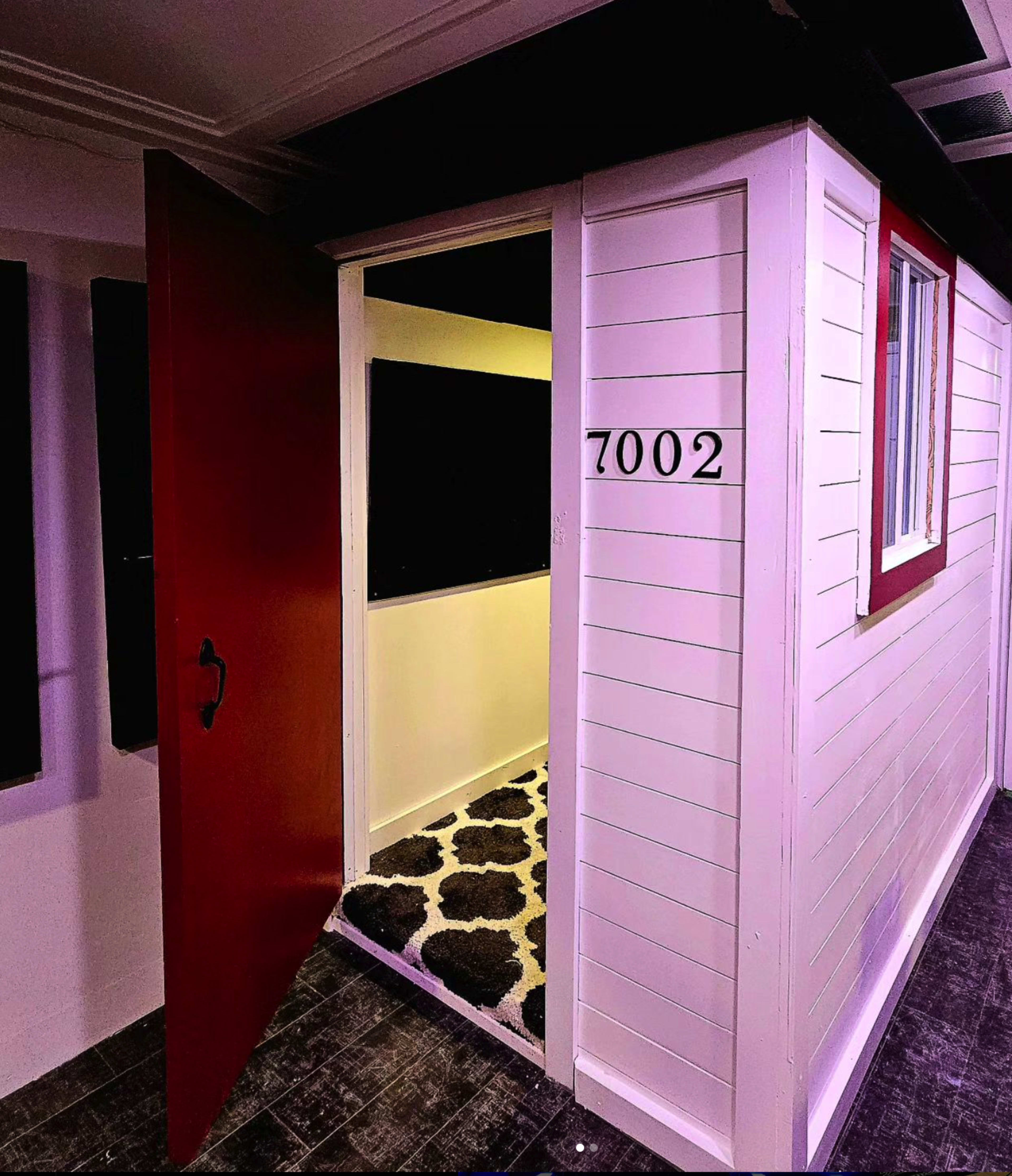 A red door opens to a bright room with the number 7002 displayed on a white exterior wall.