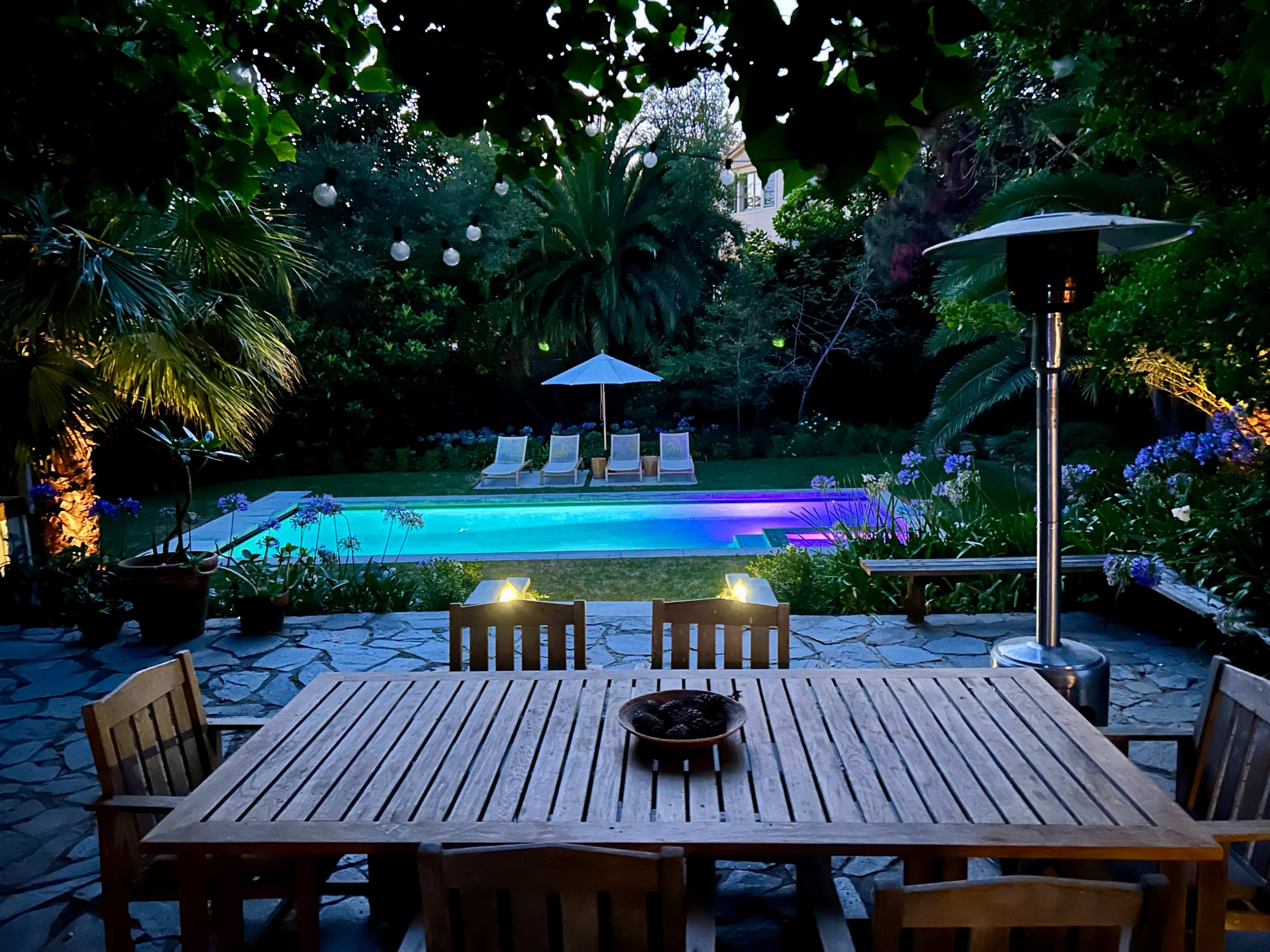 A wooden dining table is set in the foreground, overlooking a lit pool area surrounded by palm trees and lounge chairs.