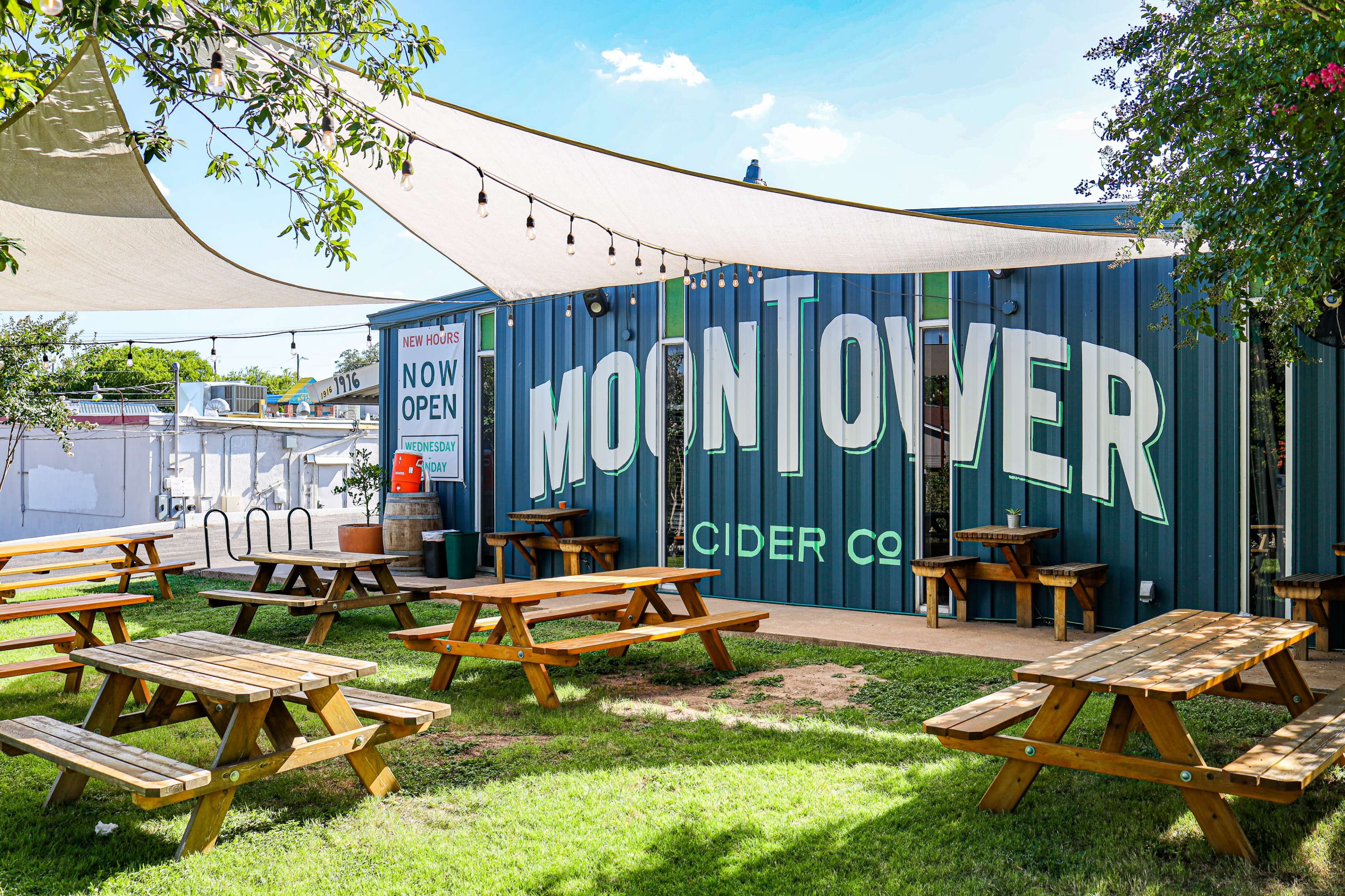 A cider company with a large mural on its side and outdoor seating made up of wooden picnic tables under shaded canopies.