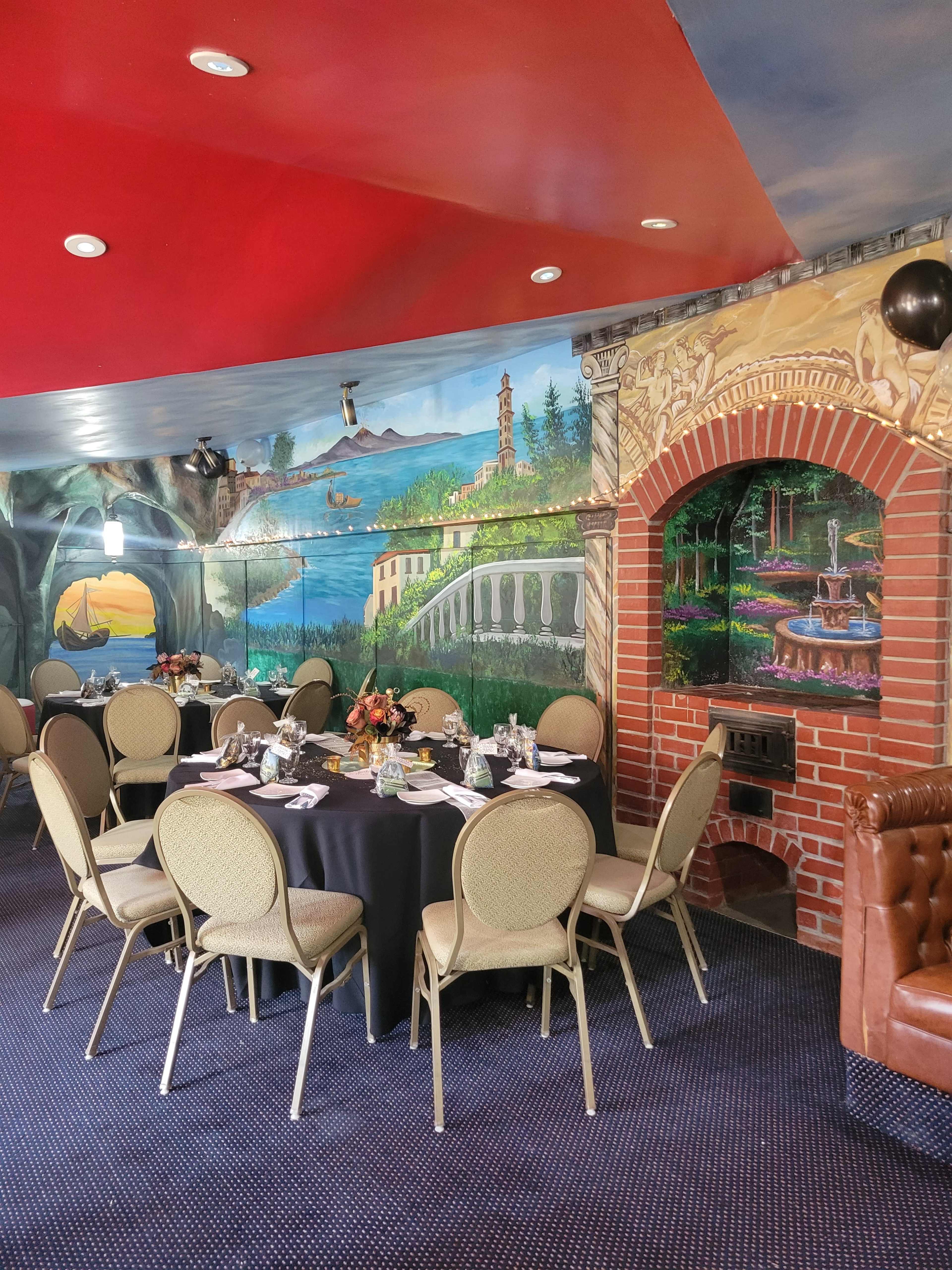 The image shows a dining area with round tables set for a meal, surrounded by a mural depicting a scenic landscape and a decorative brick archway.