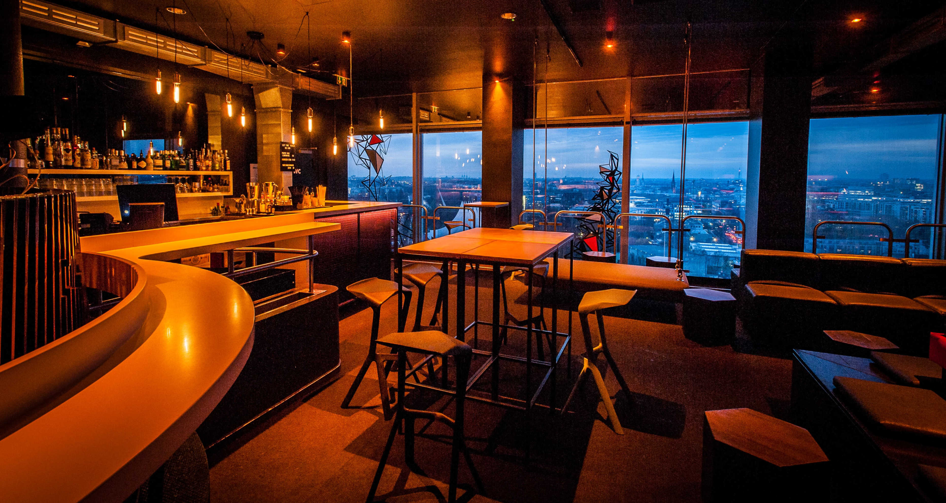 A modern bar interior with a curved countertop, high tables, and a view of the city skyline through large windows.