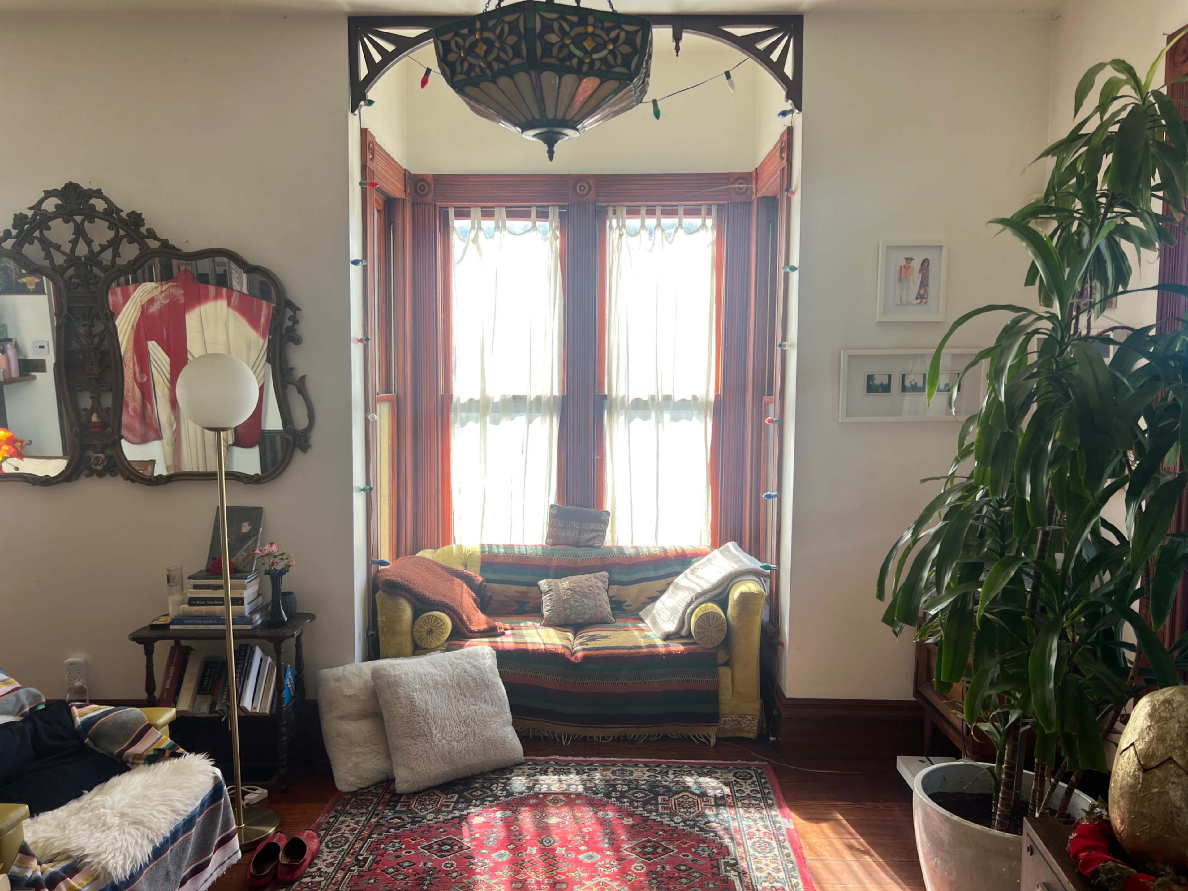 The image shows a cozy living room with a colorful couch, a large window adorned with curtains, a decorative mirror, and various plants.