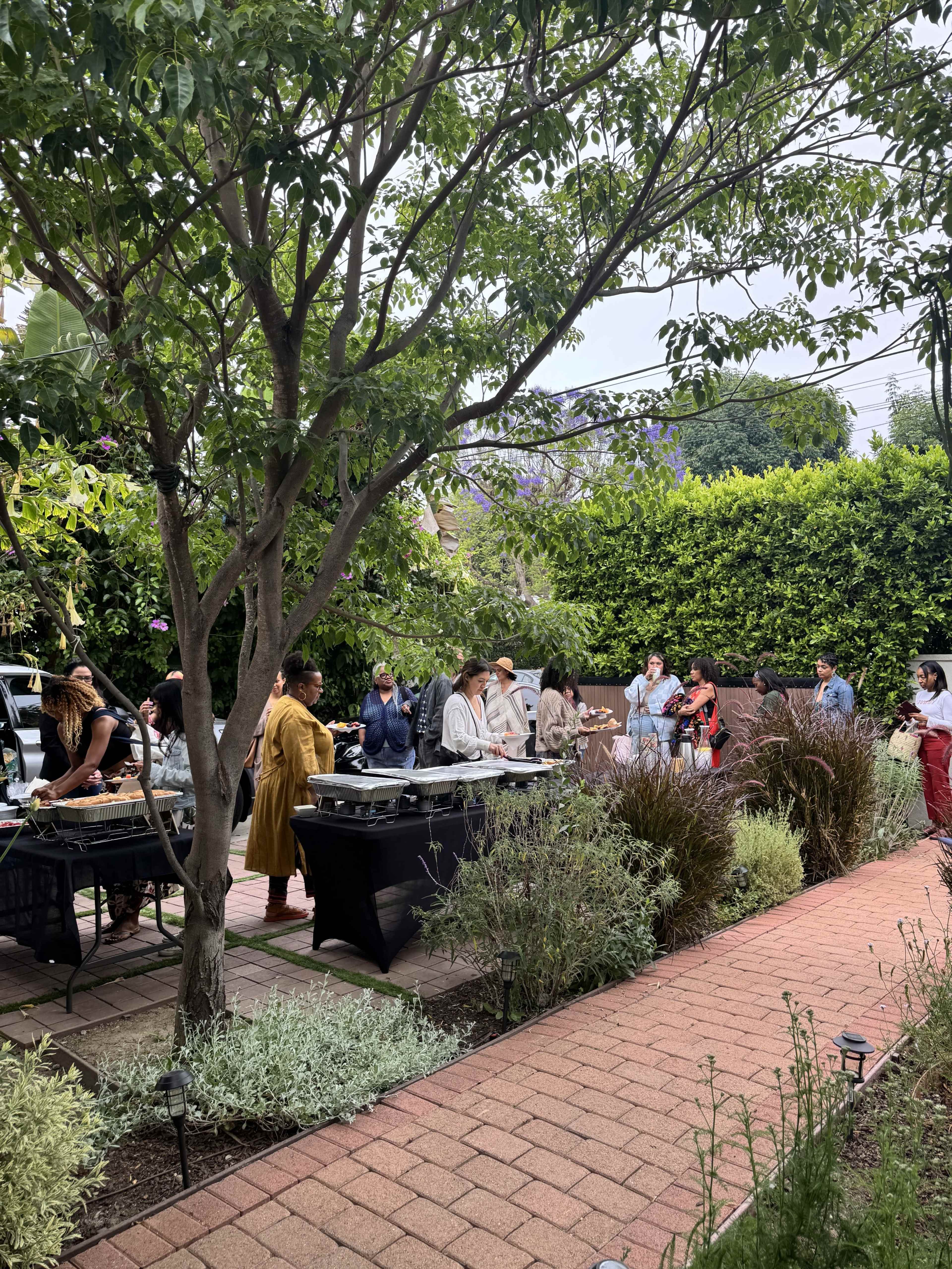 A group of people gathers in a garden for a cookout, with tables set up for food and greenery surrounding the area.