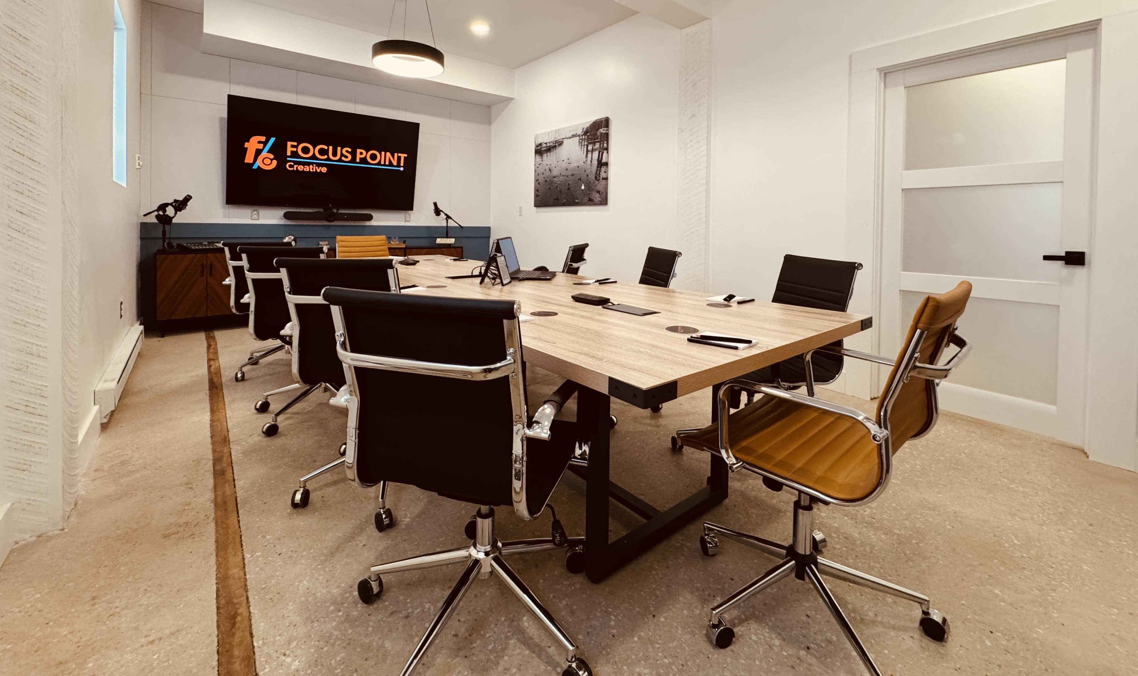 The image shows a modern conference room with a large wooden table surrounded by black and orange office chairs, featuring a wall-mounted screen displaying the "Focus Point Creative" logo.