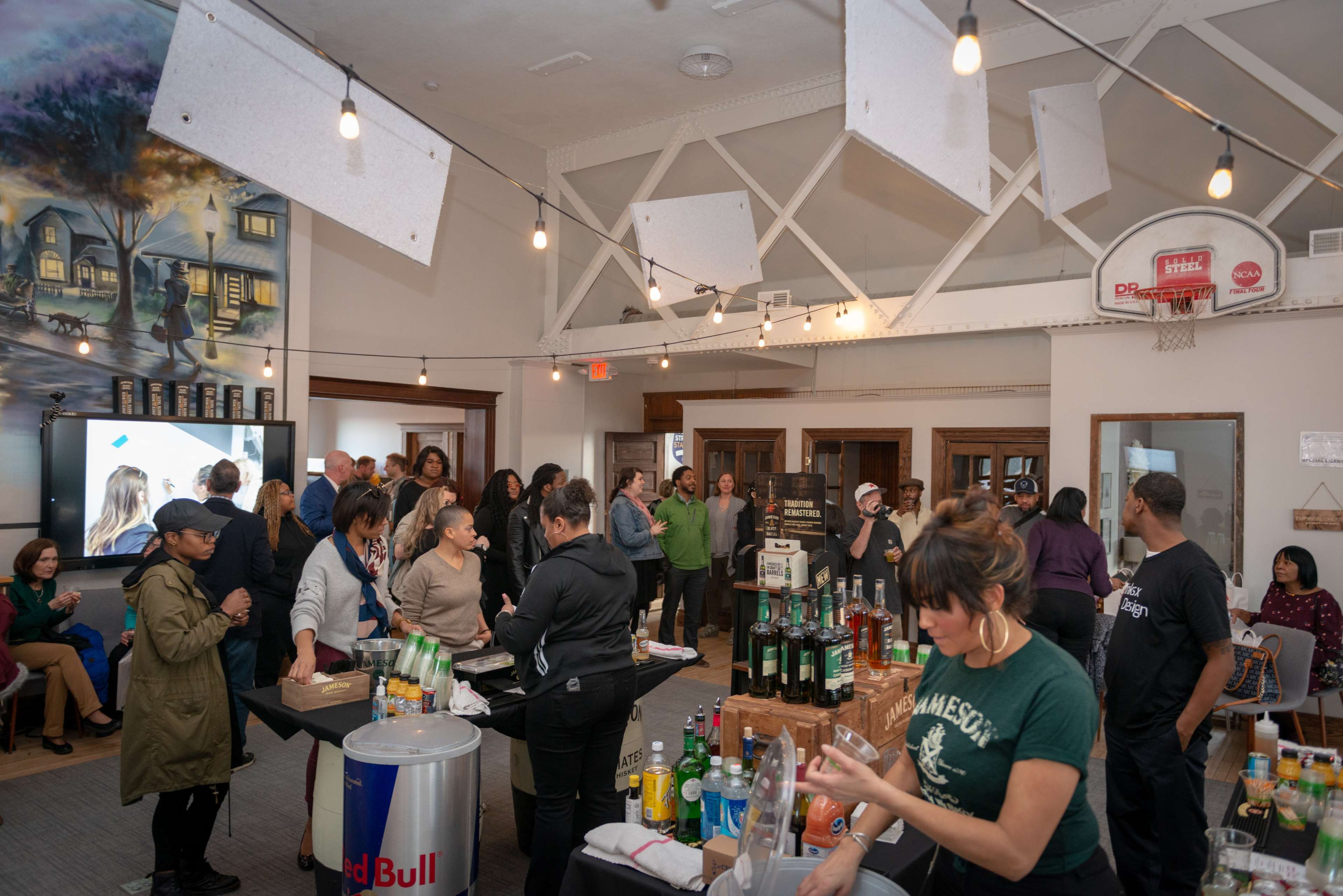 A crowd gathers in a spacious community room filled with tables displaying various beverages and a large screen showing a presentation.