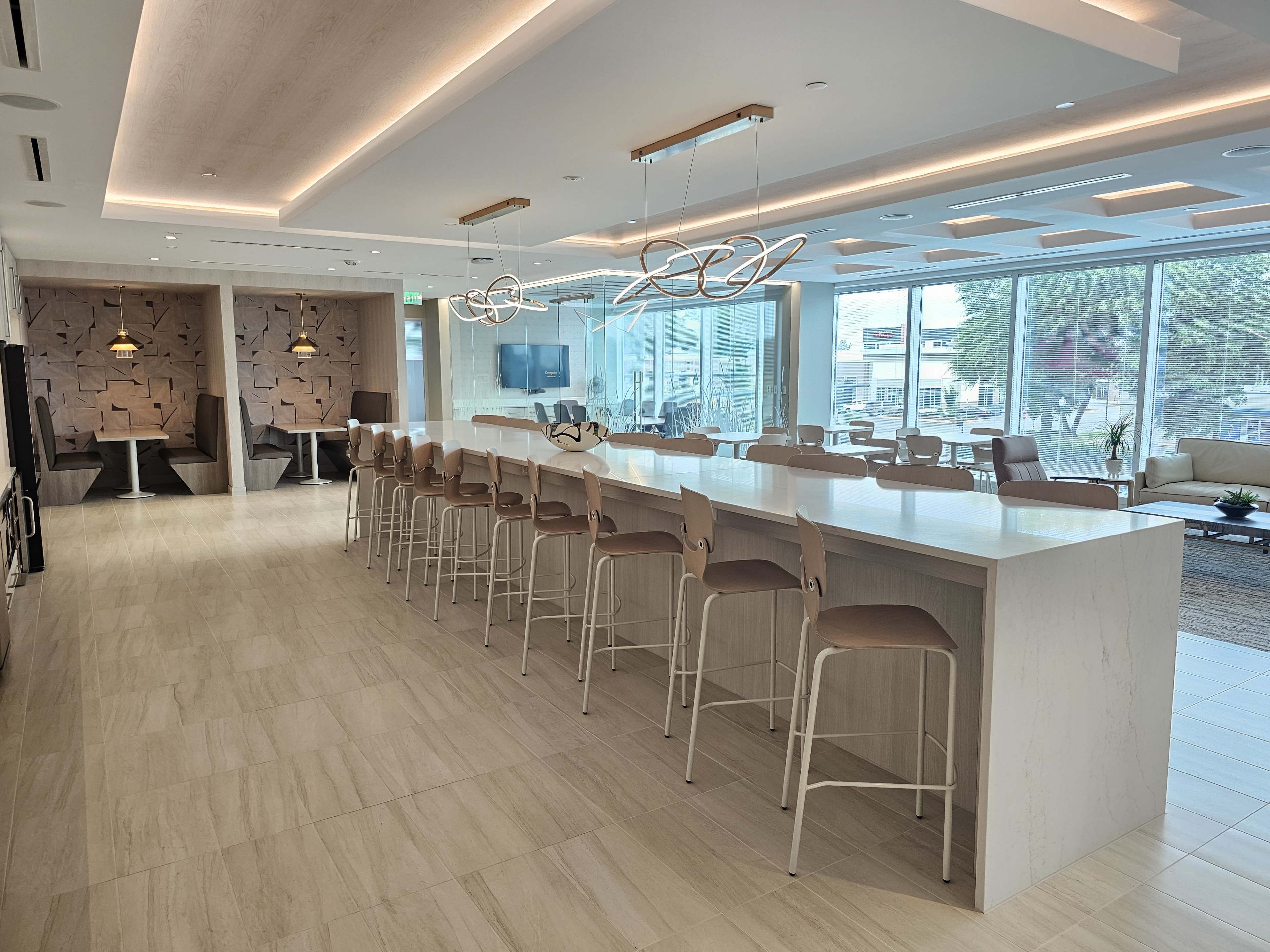 A modern, bright lounge area with a long white bar counter and high stools, surrounded by large windows.
