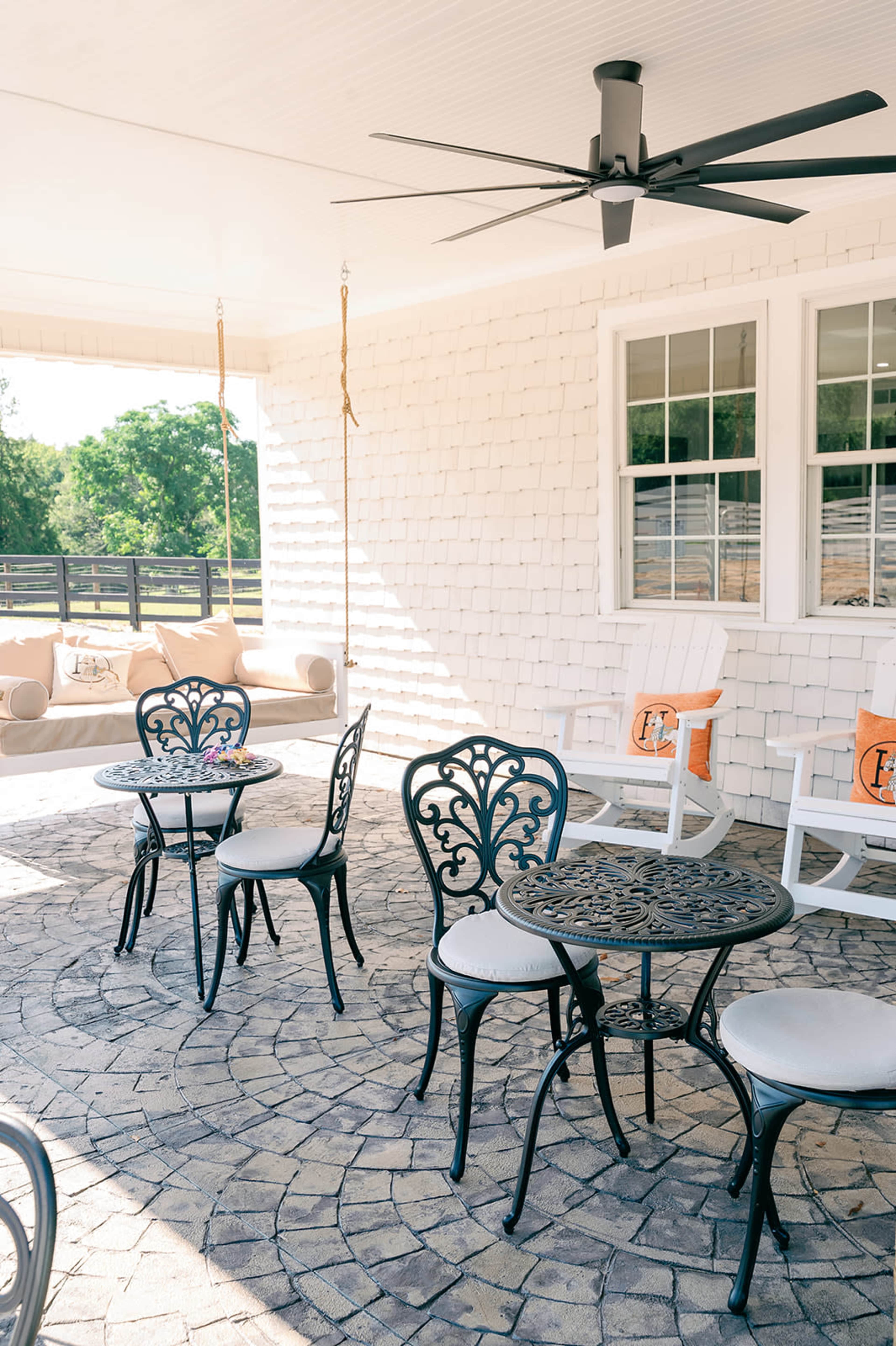 The image shows a covered patio area featuring small round tables with ornate metal chairs, a ceiling fan, and a seating area with cushions in the background.