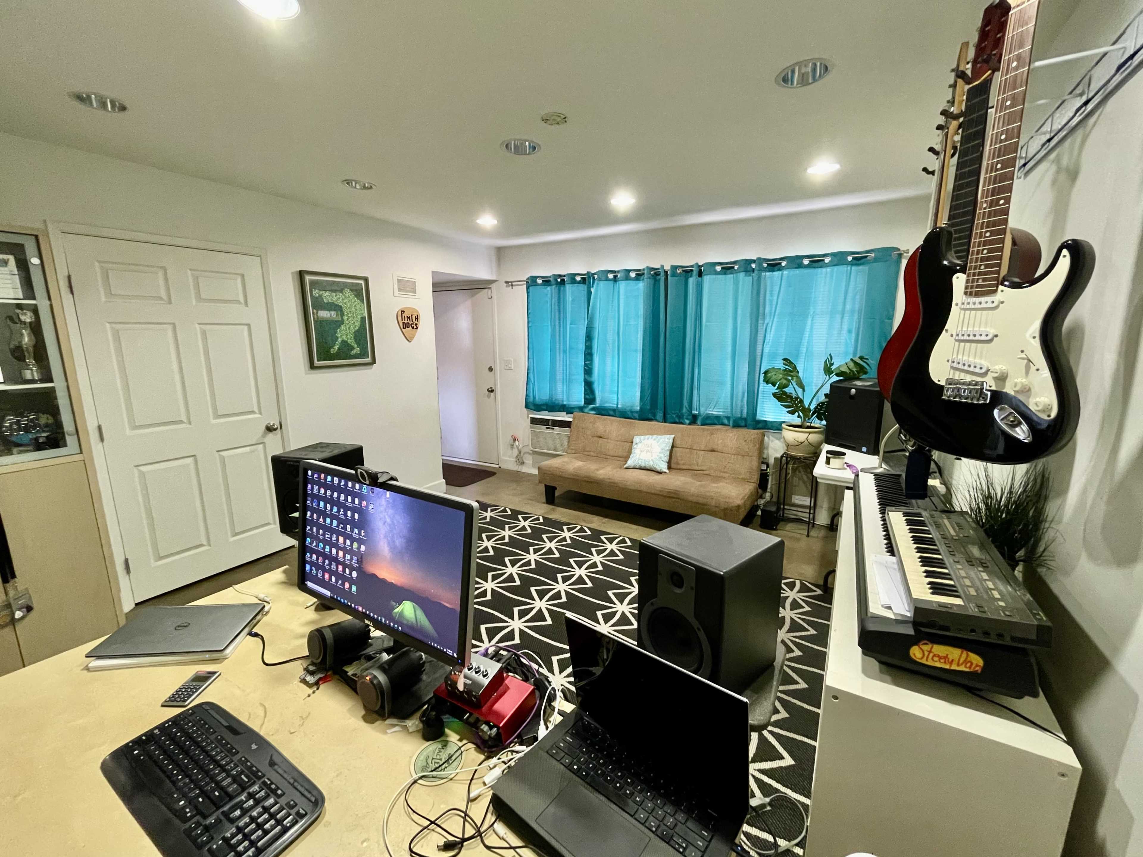 The image shows a home office space featuring a desk with a computer, a laptop, a keyboard, and a guitar hanging on the wall, along with a sofa and curtains in the background.