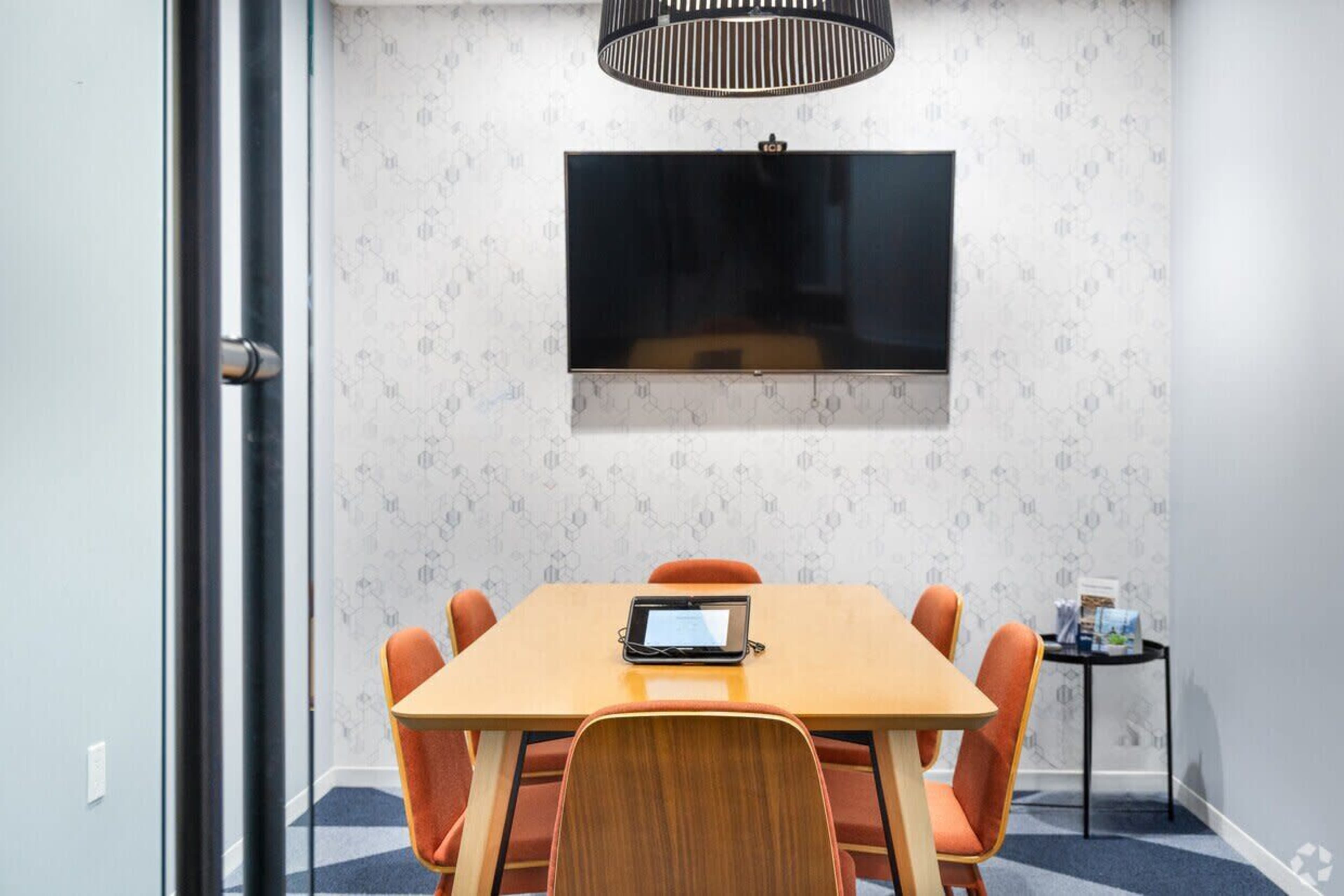 A conference room features a wooden table surrounded by orange chairs, with a large flat-screen TV mounted on the wall.