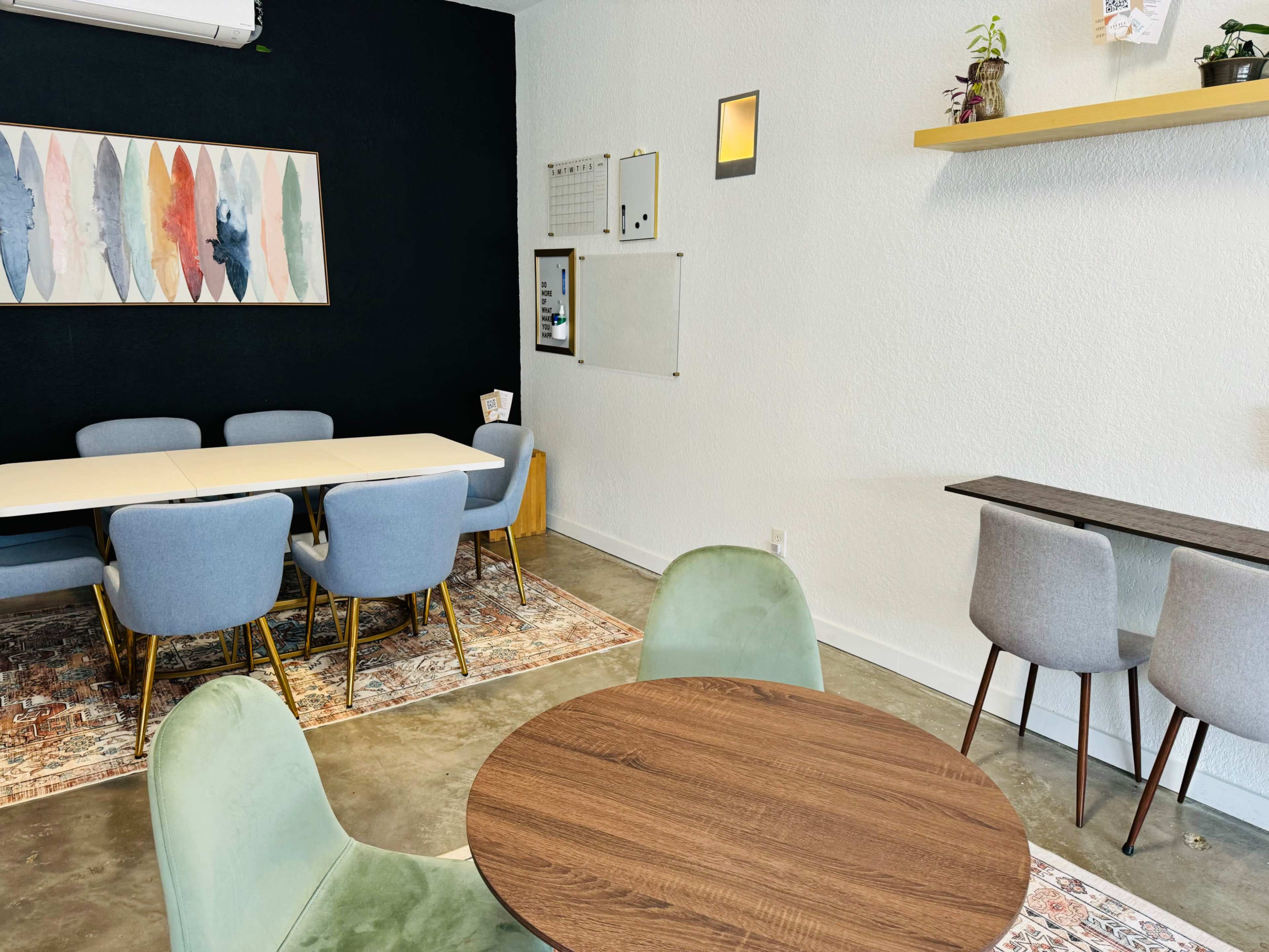 A modern, minimalistic interior with a dining area featuring a long table surrounded by blue chairs, a circular wooden table, and a wall with decorative artwork and plants.