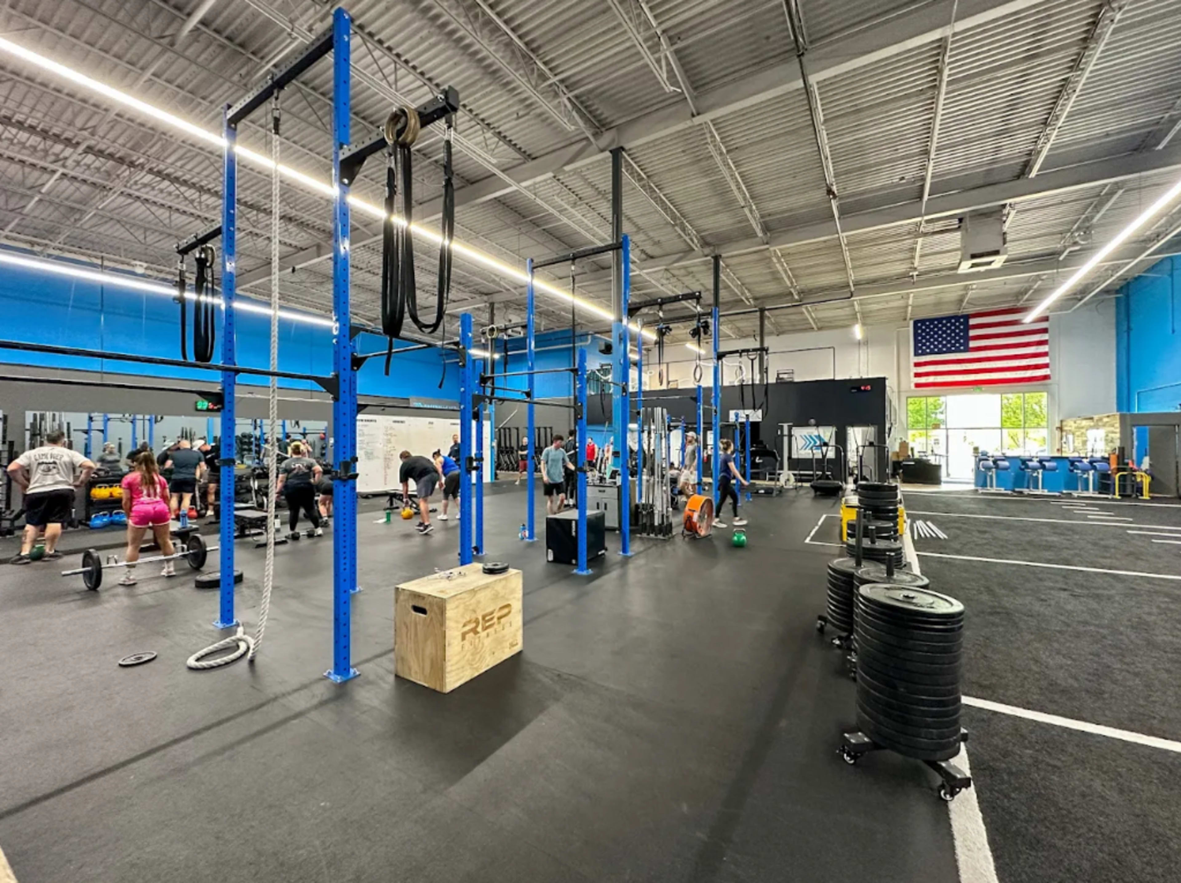 The image shows a gym with workout equipment, various fitness stations, and individuals engaged in exercise activities, accompanied by an American flag on the wall.