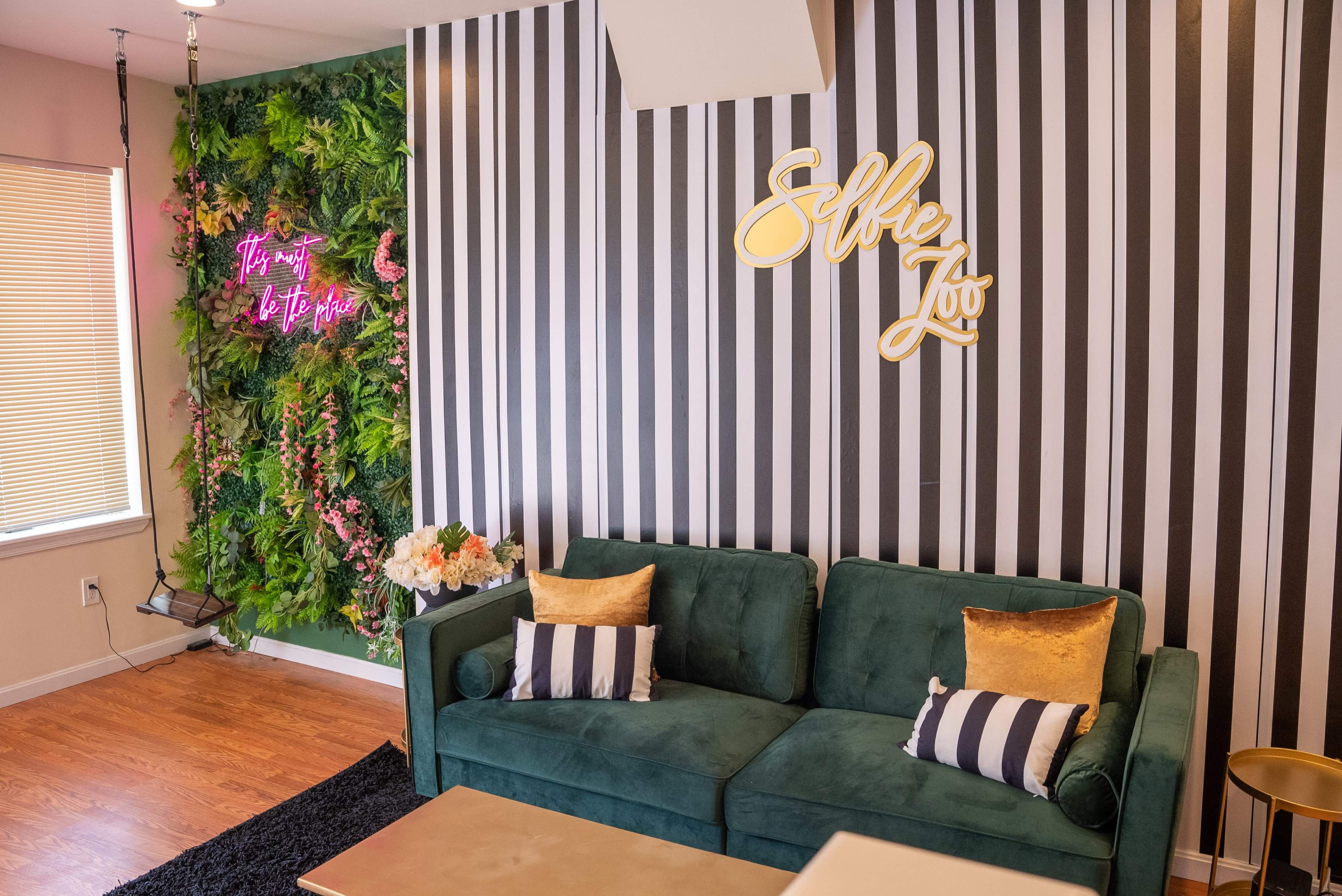 The image shows a modern interior with a green velvet sofa, striped wallpaper, and a lively green wall adorned with plants and a neon sign that reads "Selfie Zoo."