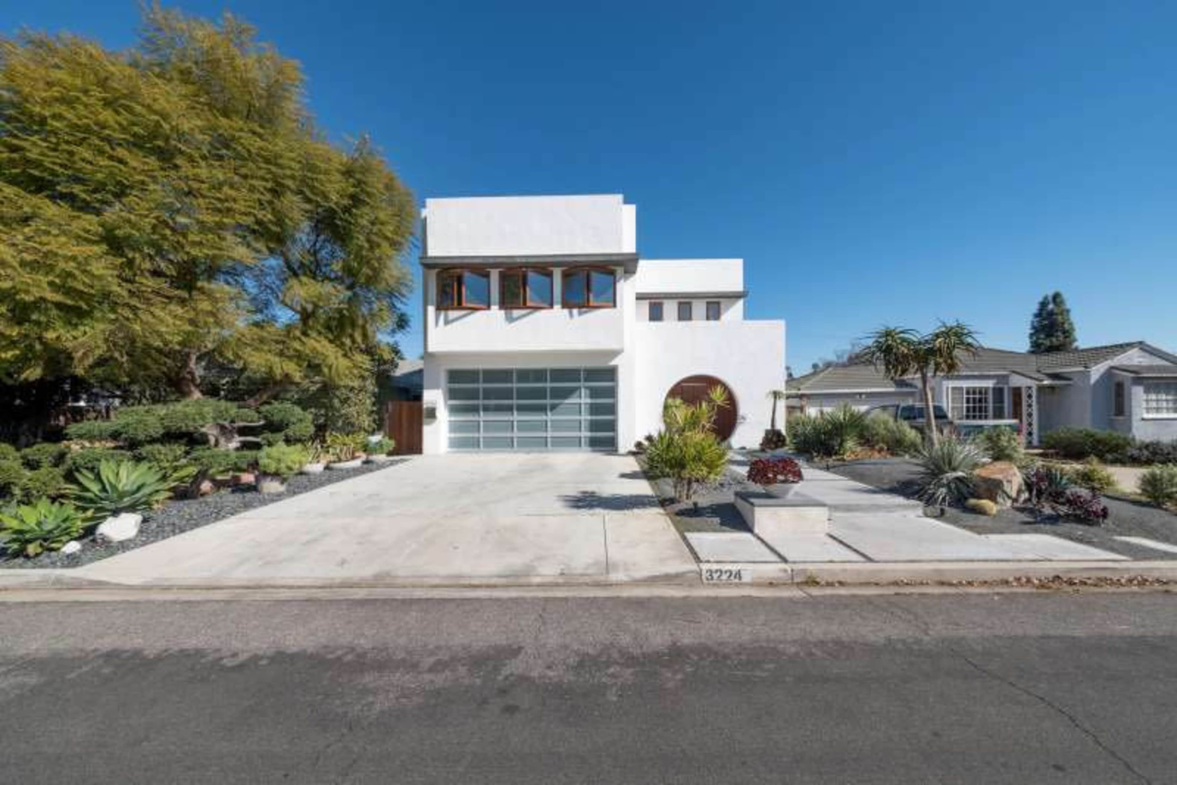 A modern two-story white house with large windows and a spacious front driveway, surrounded by landscaped greenery.