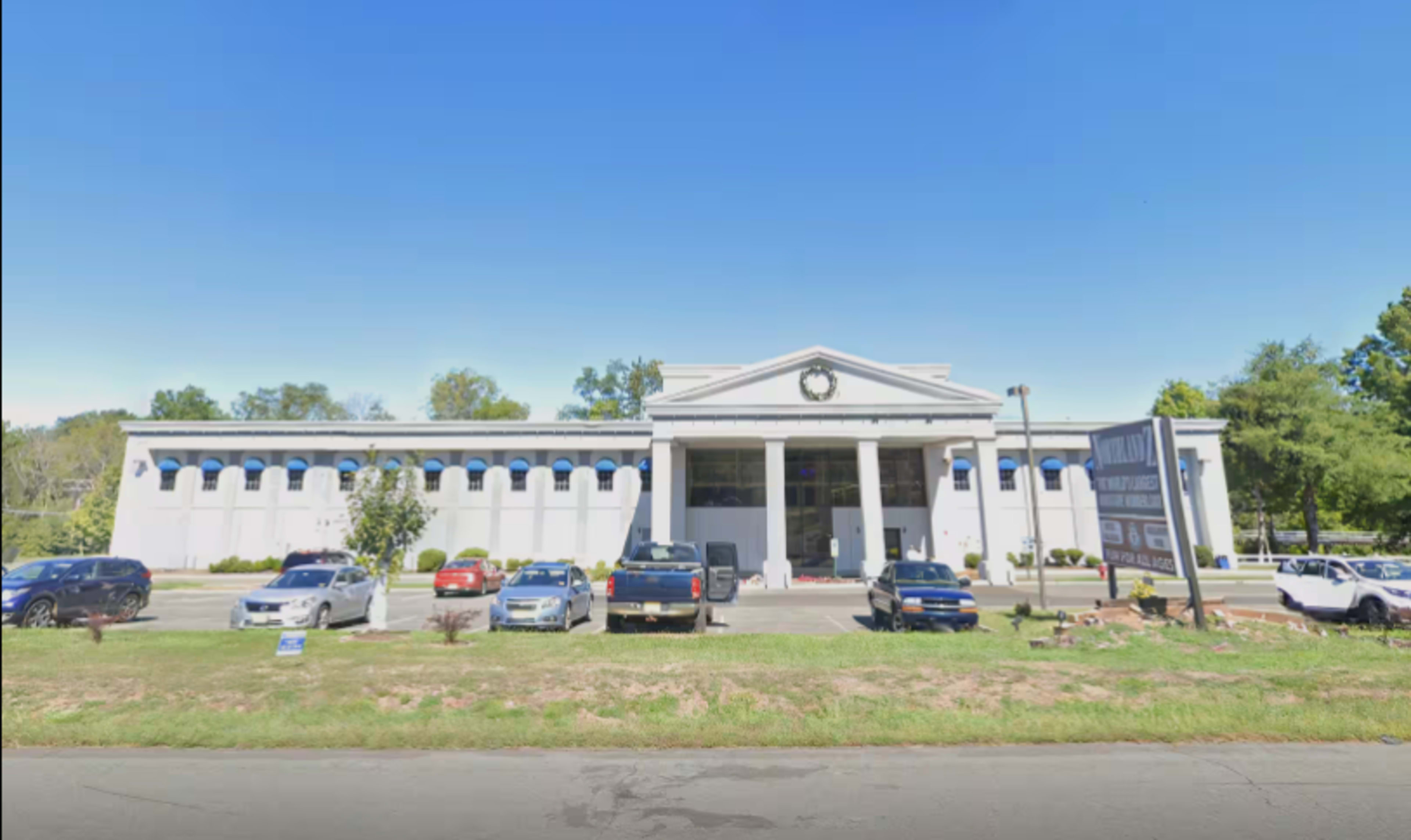 The image shows a large, white building with a columned entrance and multiple vehicles parked in front.