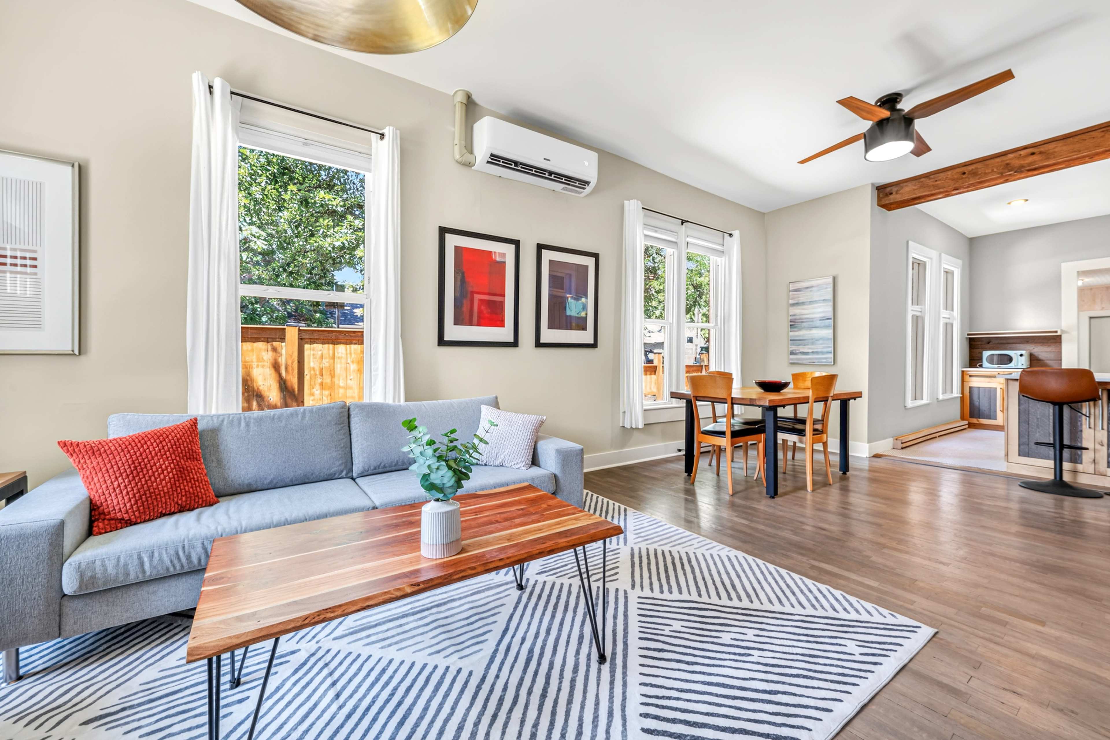 The image shows a bright, modern living space featuring a gray sofa, a wooden coffee table, and a dining area with a table and chairs, illuminated by natural light through large windows.