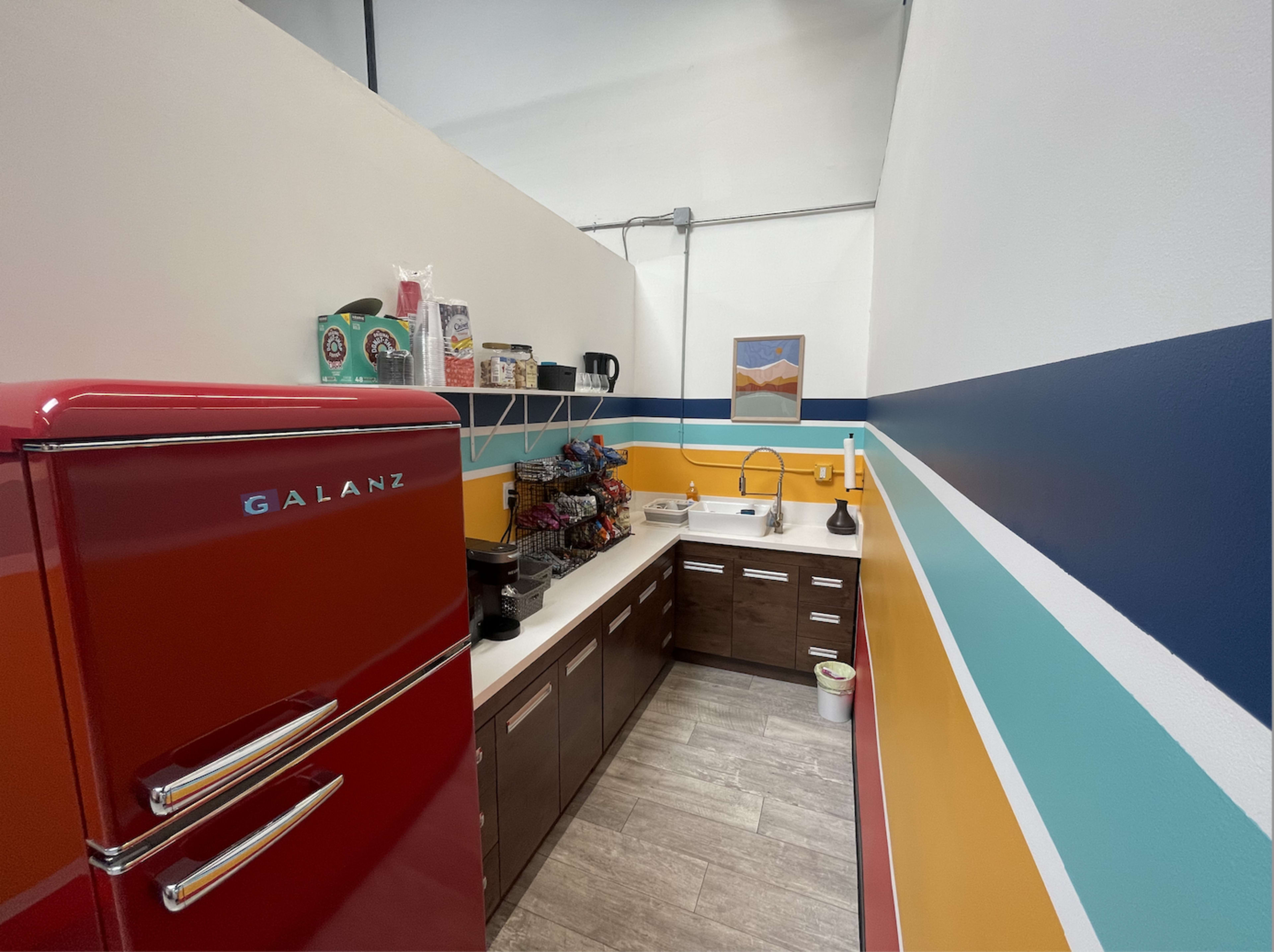 A compact kitchen features a red refrigerator, a sink, and cabinets against colorful striped walls.
