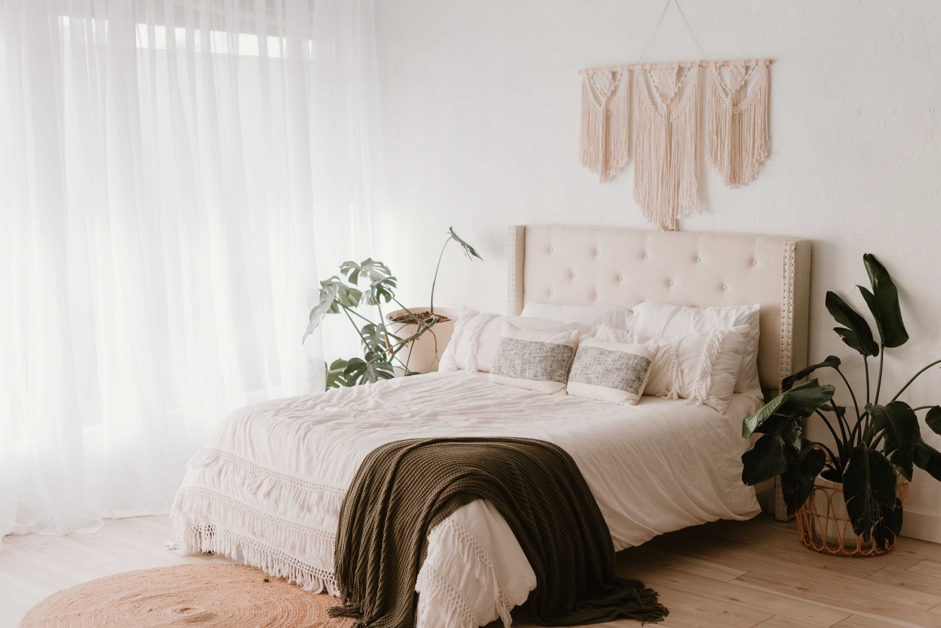 A neatly made bed with a decorative headboard, surrounded by indoor plants and bathed in natural light from large sheer curtains.