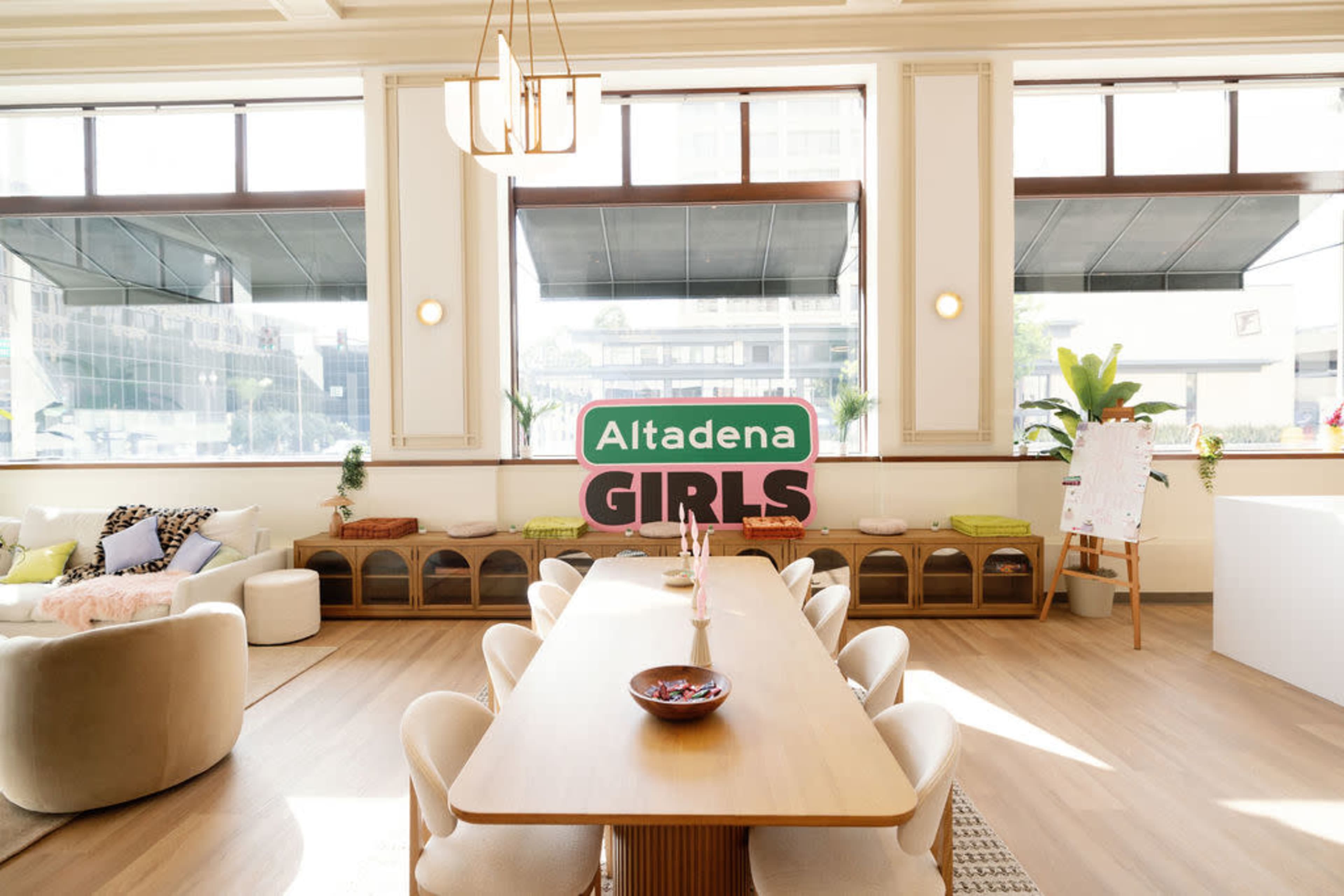 The image shows a modern, well-lit interior of a community space featuring a long dining table, comfortable seating areas, and large windows.