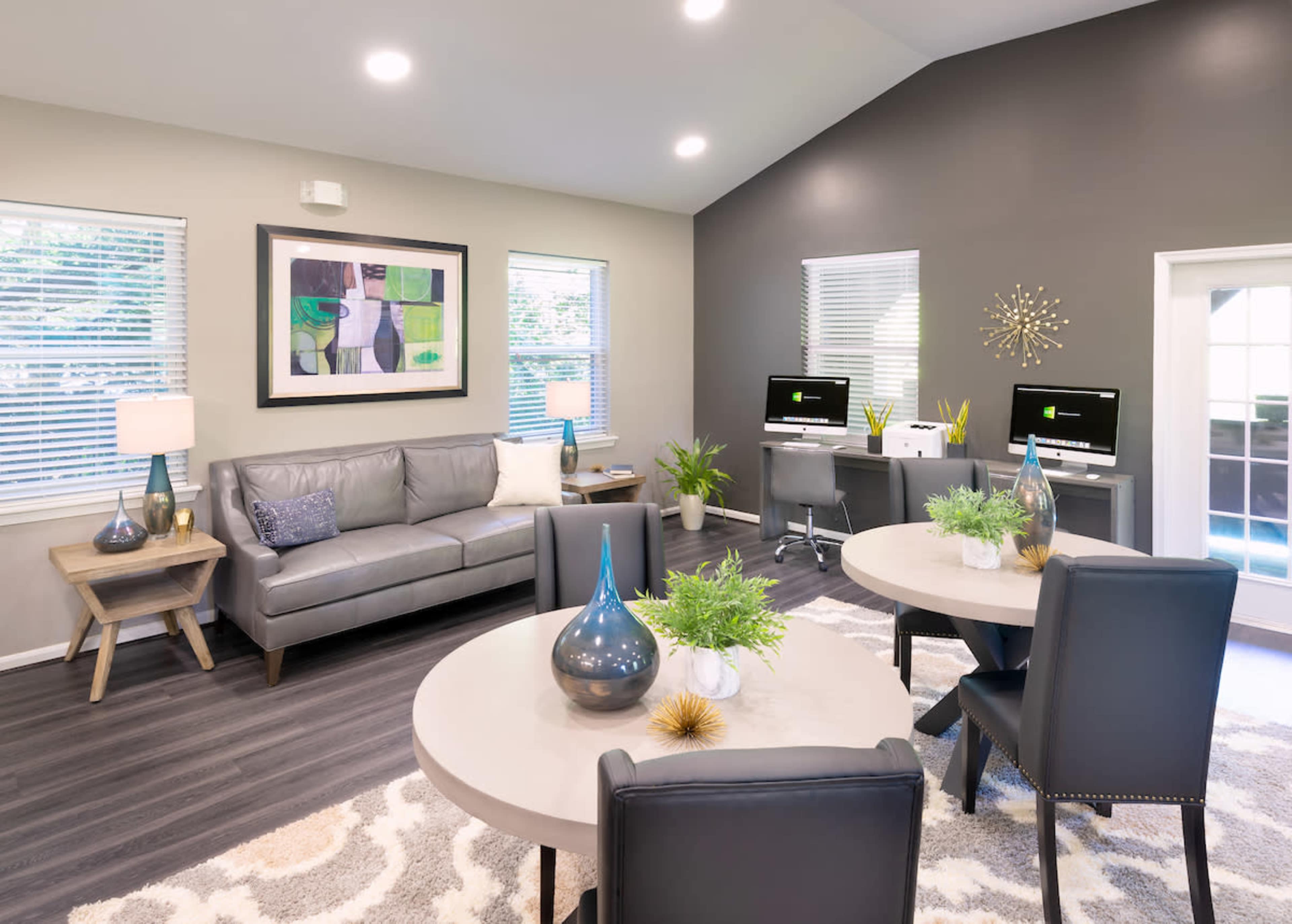 A modern living space features a gray sofa, two round tables, and workstations with computers, alongside large windows allowing for natural light.