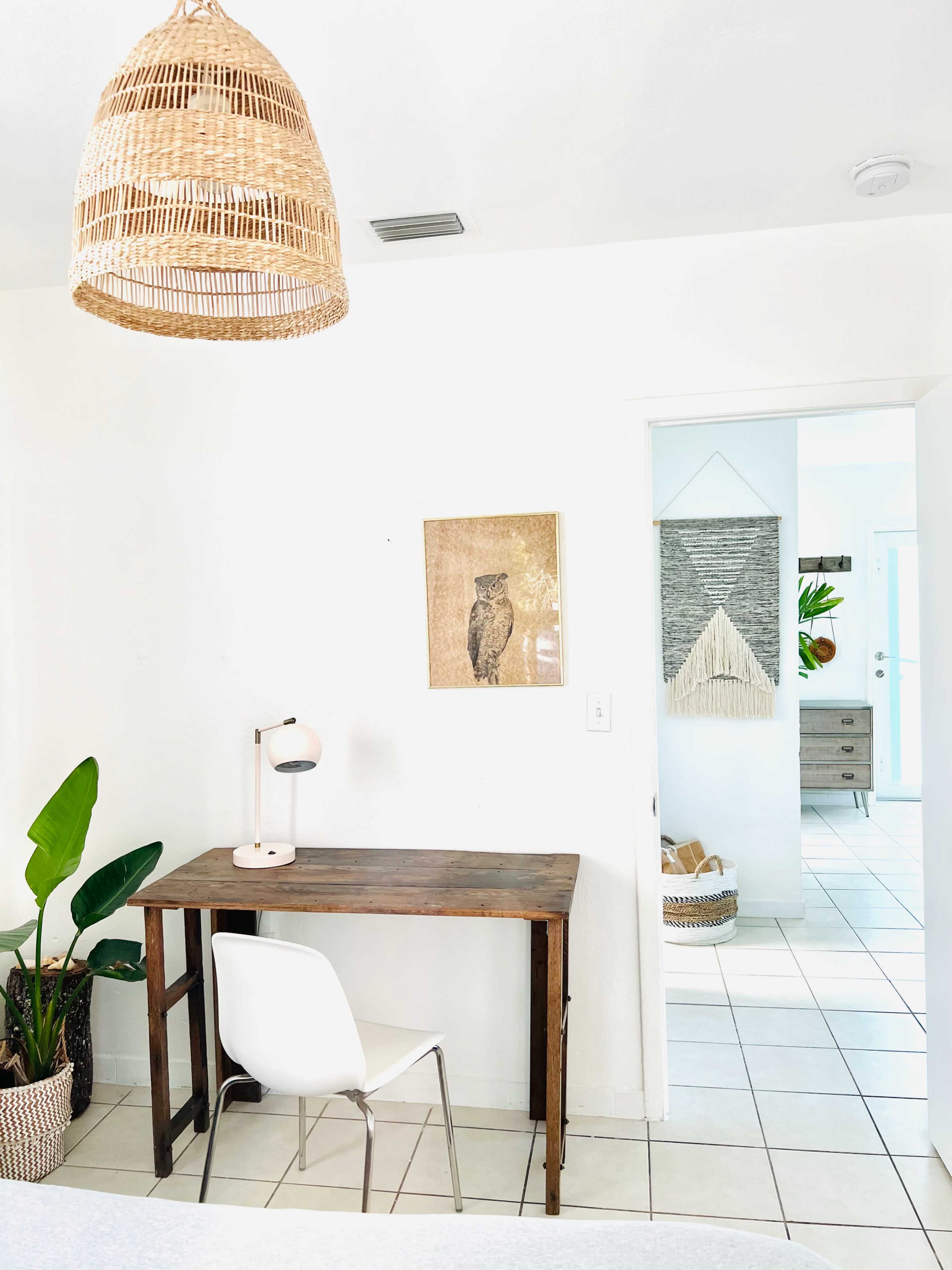A wooden desk and white chair are positioned against a wall adorned with an owl print, with a large pendant light hanging above in a well-lit room.