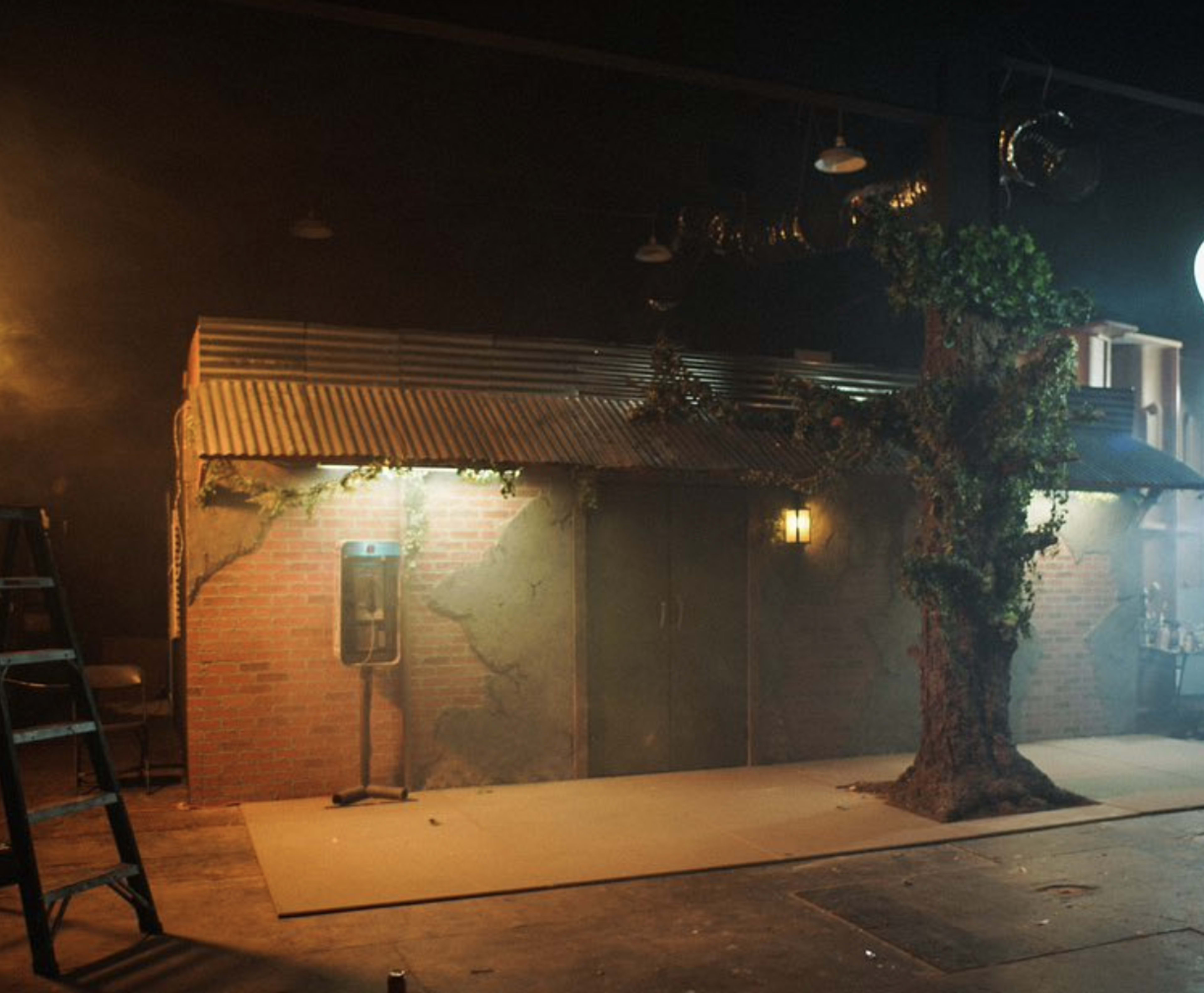 A small, rustic building with a corrugated metal roof, surrounded by fog and a tree, set in a dimly lit warehouse space.