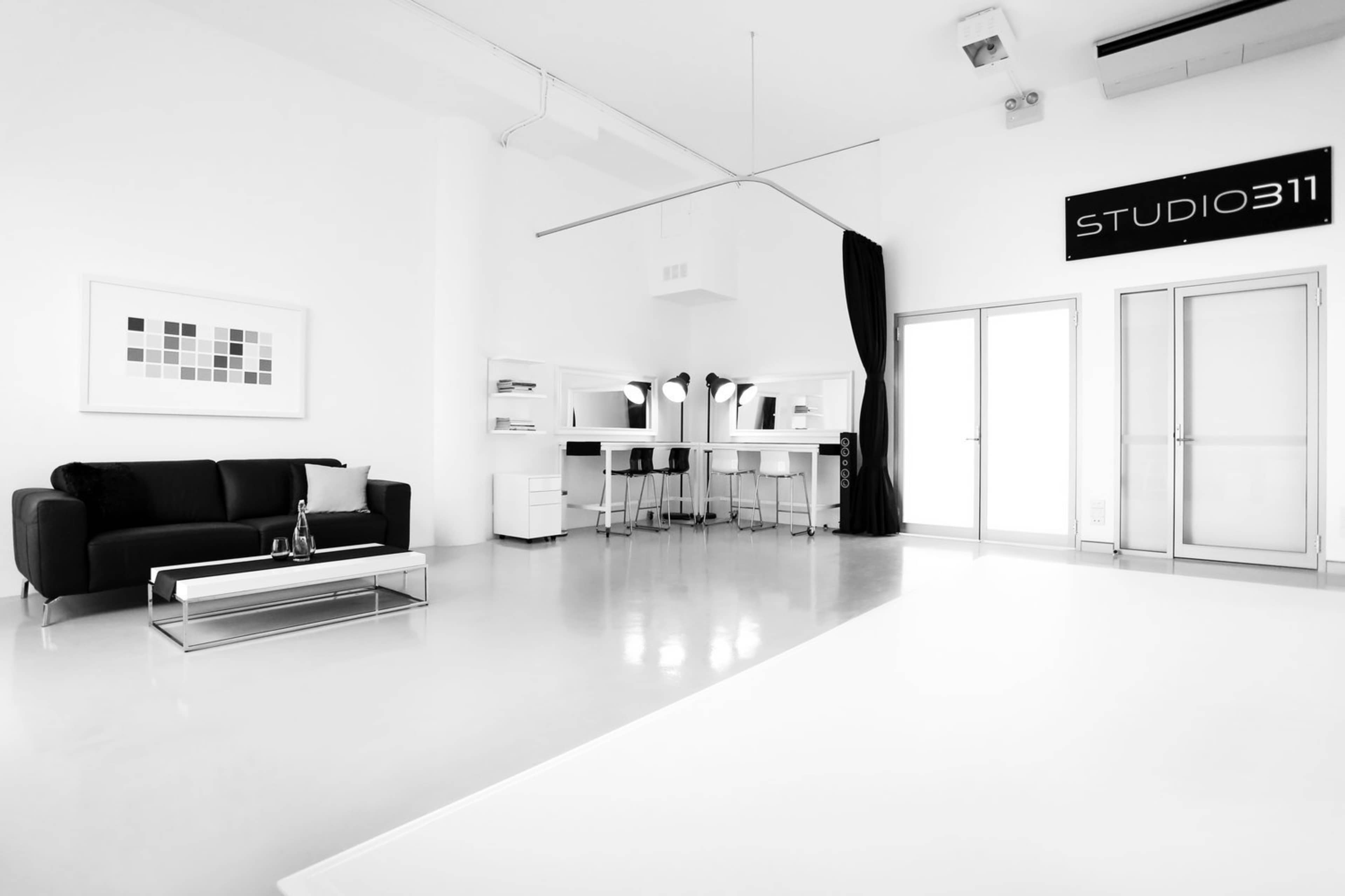 A minimalist studio space features a black sofa, a white coffee table, and two makeup stations beside a large window.