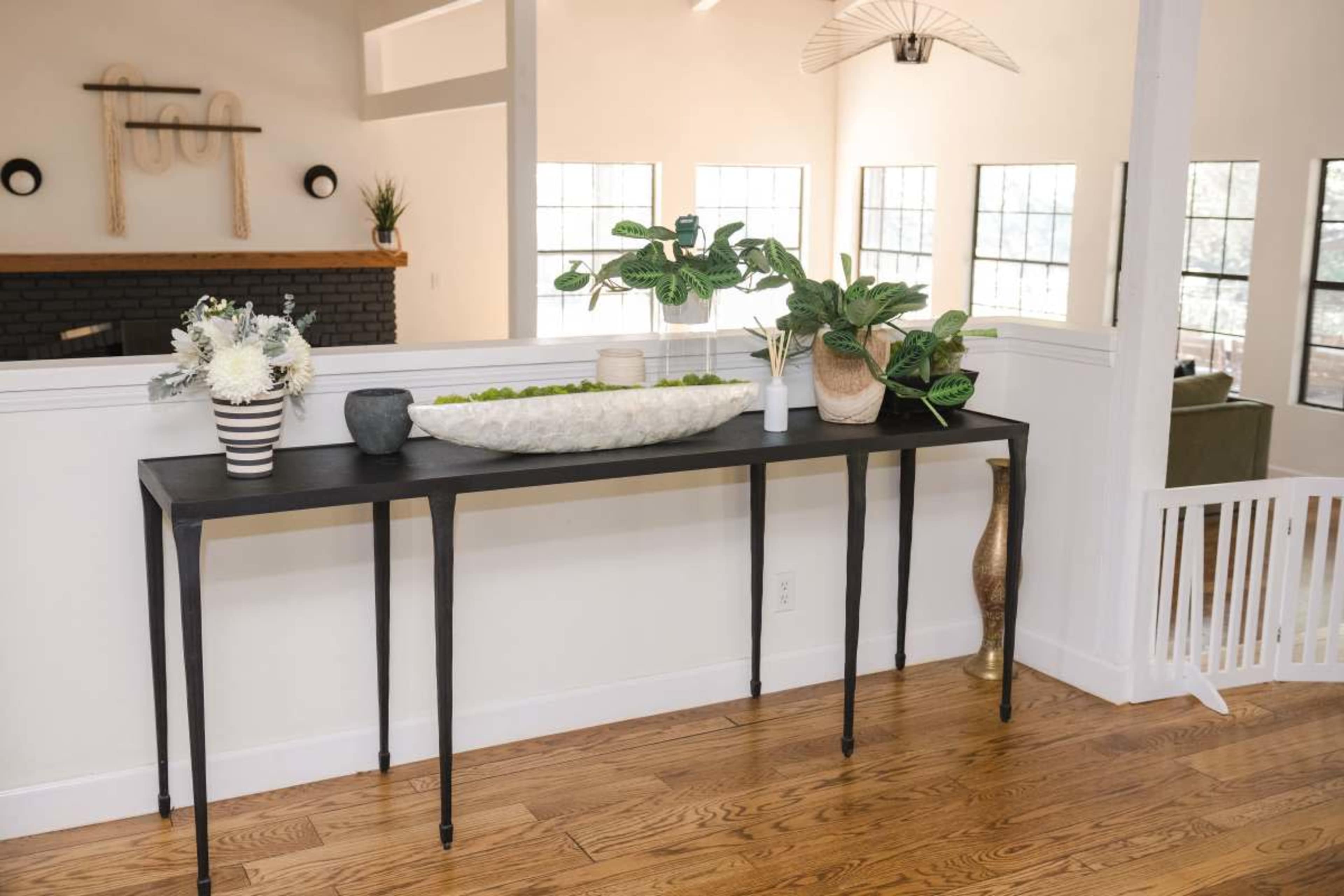 A long black console table with elegant legs displays a decorative bowl filled with greenery and is adorned with potted plants and a vase of flowers.