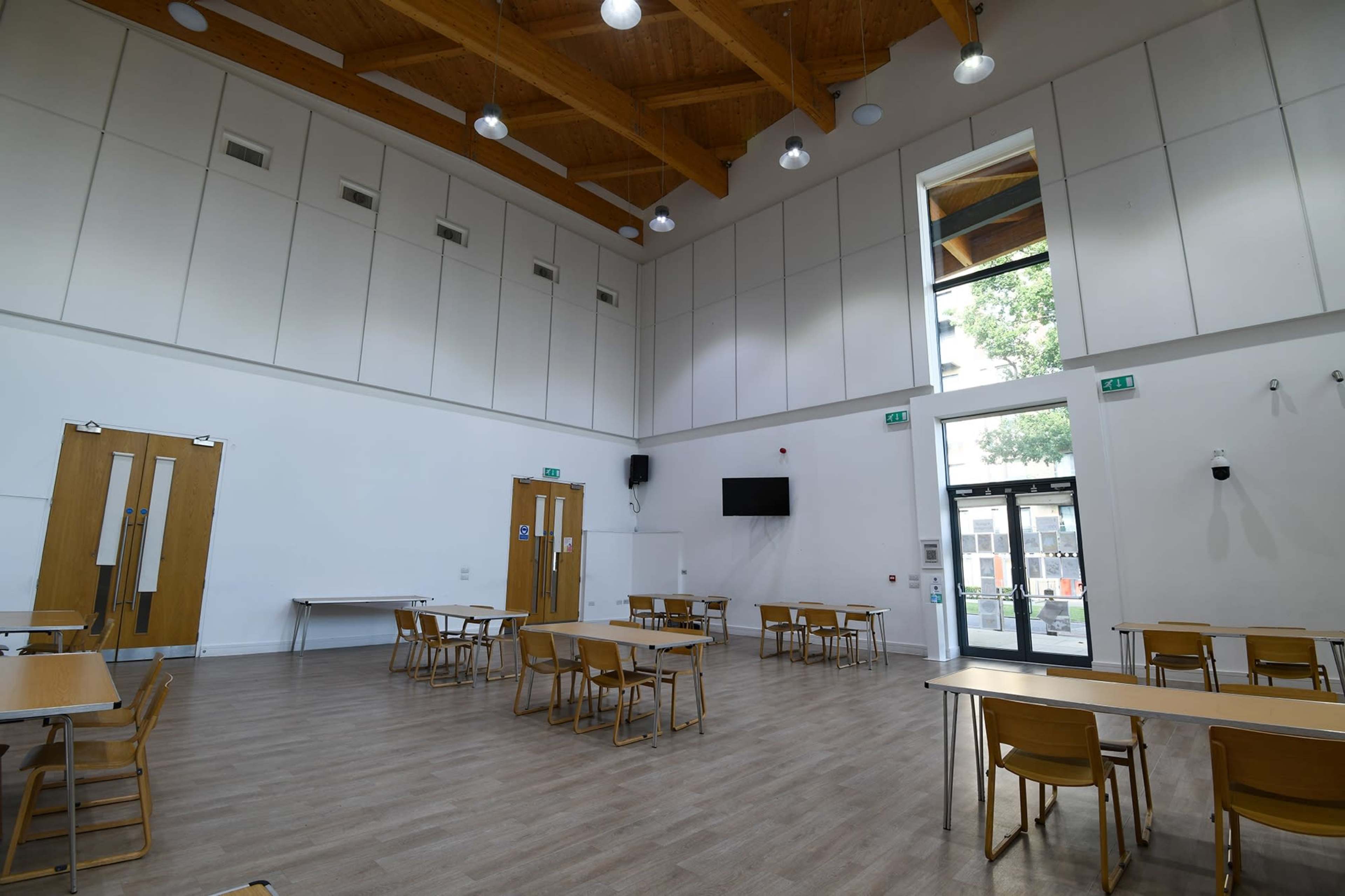 A spacious, well-lit room with wooden beams, several tables and chairs arranged neatly, and large windows letting in natural light.