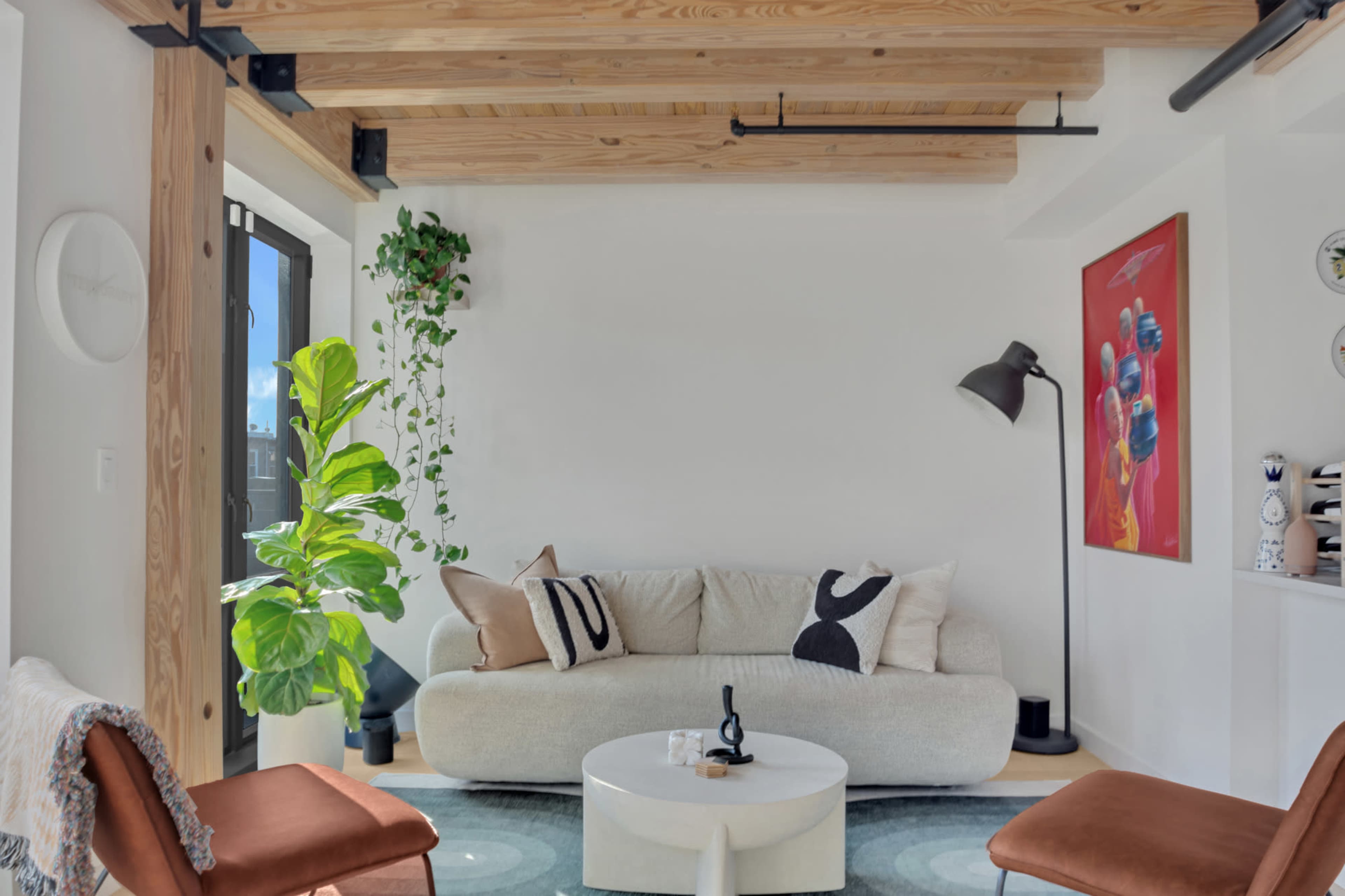 The image shows a modern living room with a curved sofa, a round coffee table, a floor lamp, and a large plant, complemented by wood-beamed ceilings and colorful artwork on the walls.