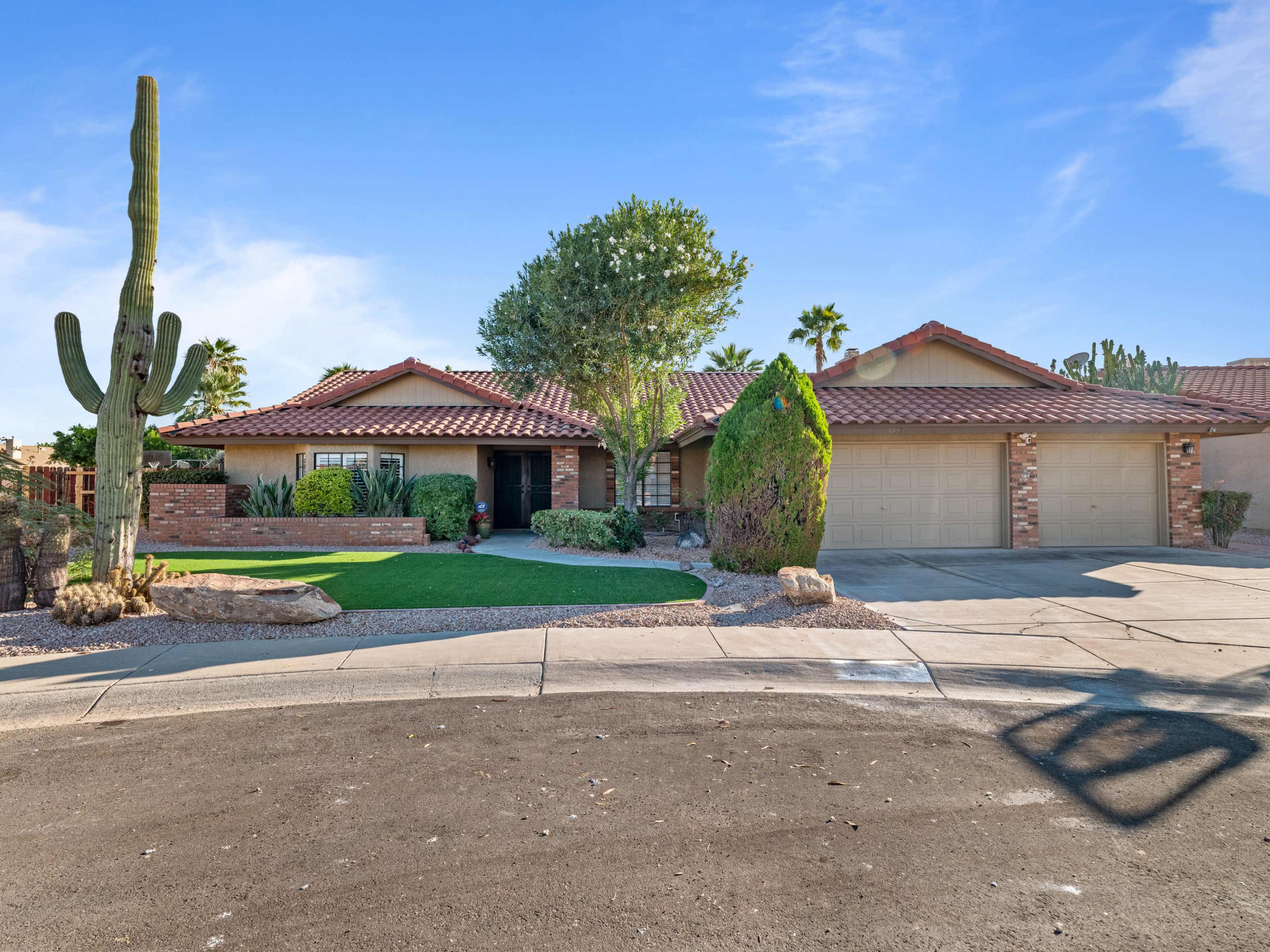 A single-story house with a red tile roof and two garage doors is situated on a corner lot, surrounded by landscaped grass and desert plants.