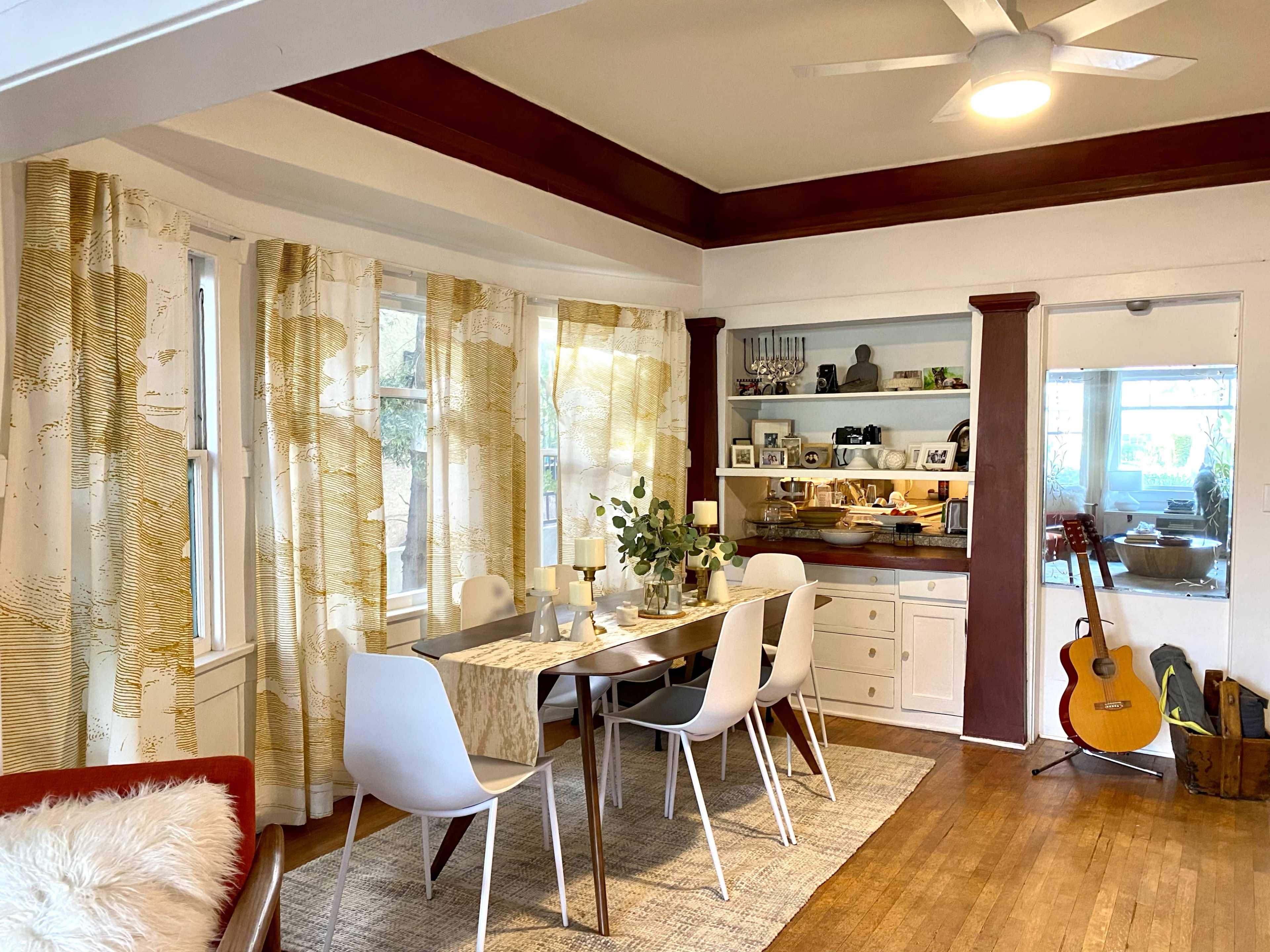 A dining area features a table set for meals, surrounded by white chairs, with curtains, a guitar, and shelves displaying decorative items.