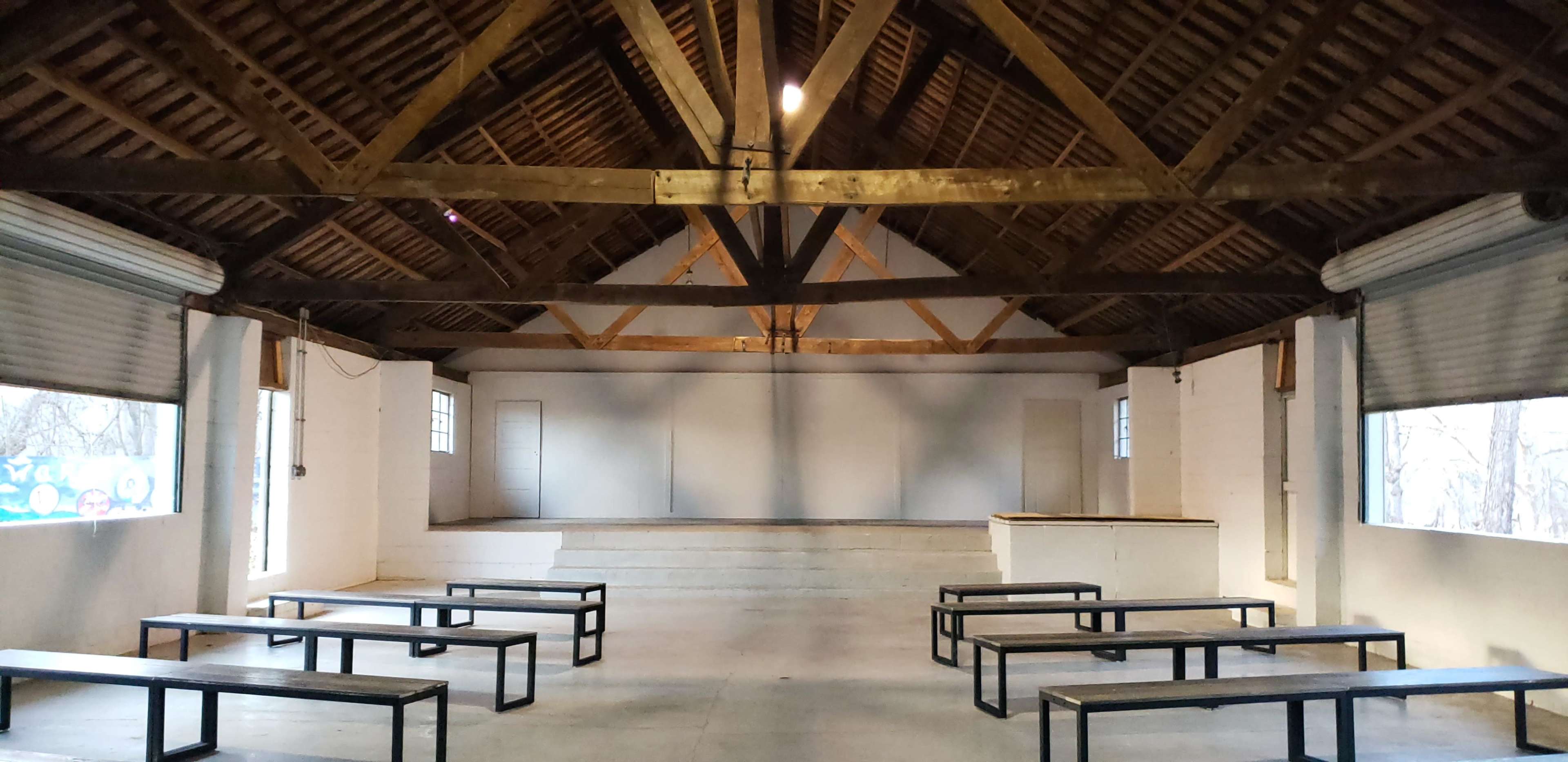 A spacious indoor venue with a high wooden ceiling, featuring benches arranged in front of a raised stage and two doors on the side walls.