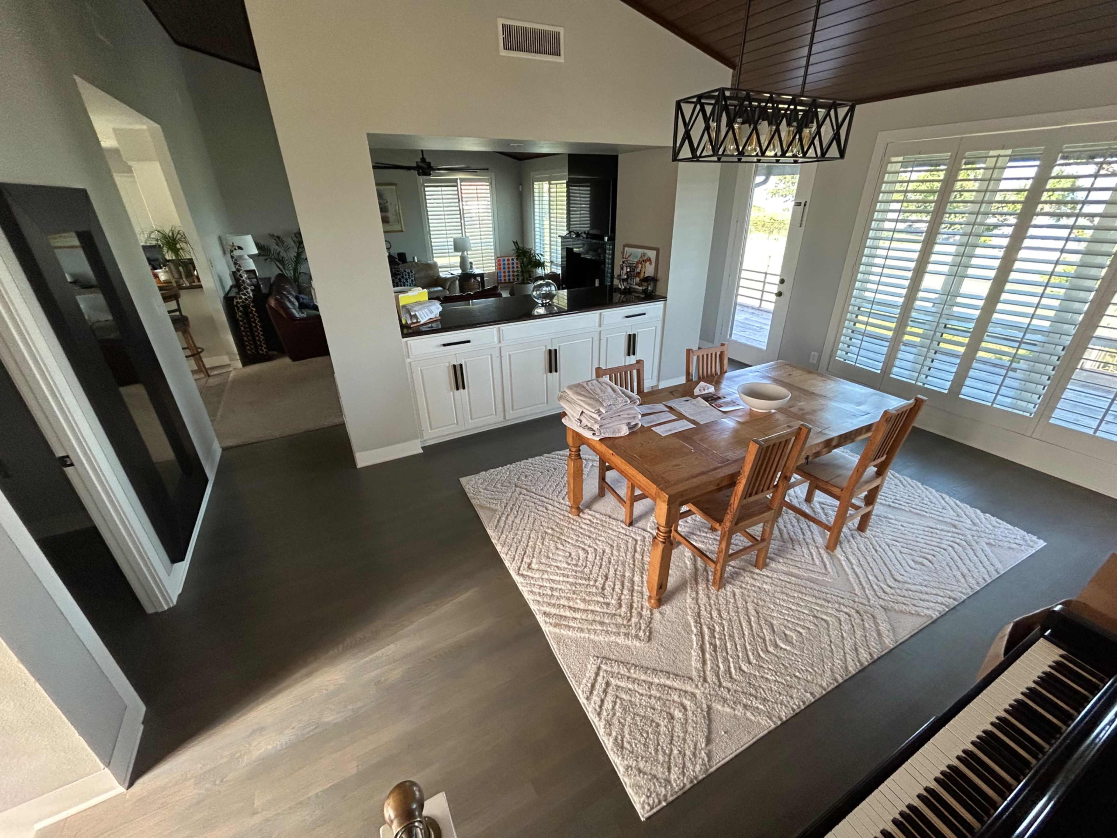 The image shows a well-lit dining area with a wooden table surrounded by chairs, a patterned rug on the floor, and large windows allowing natural light to enter the space.