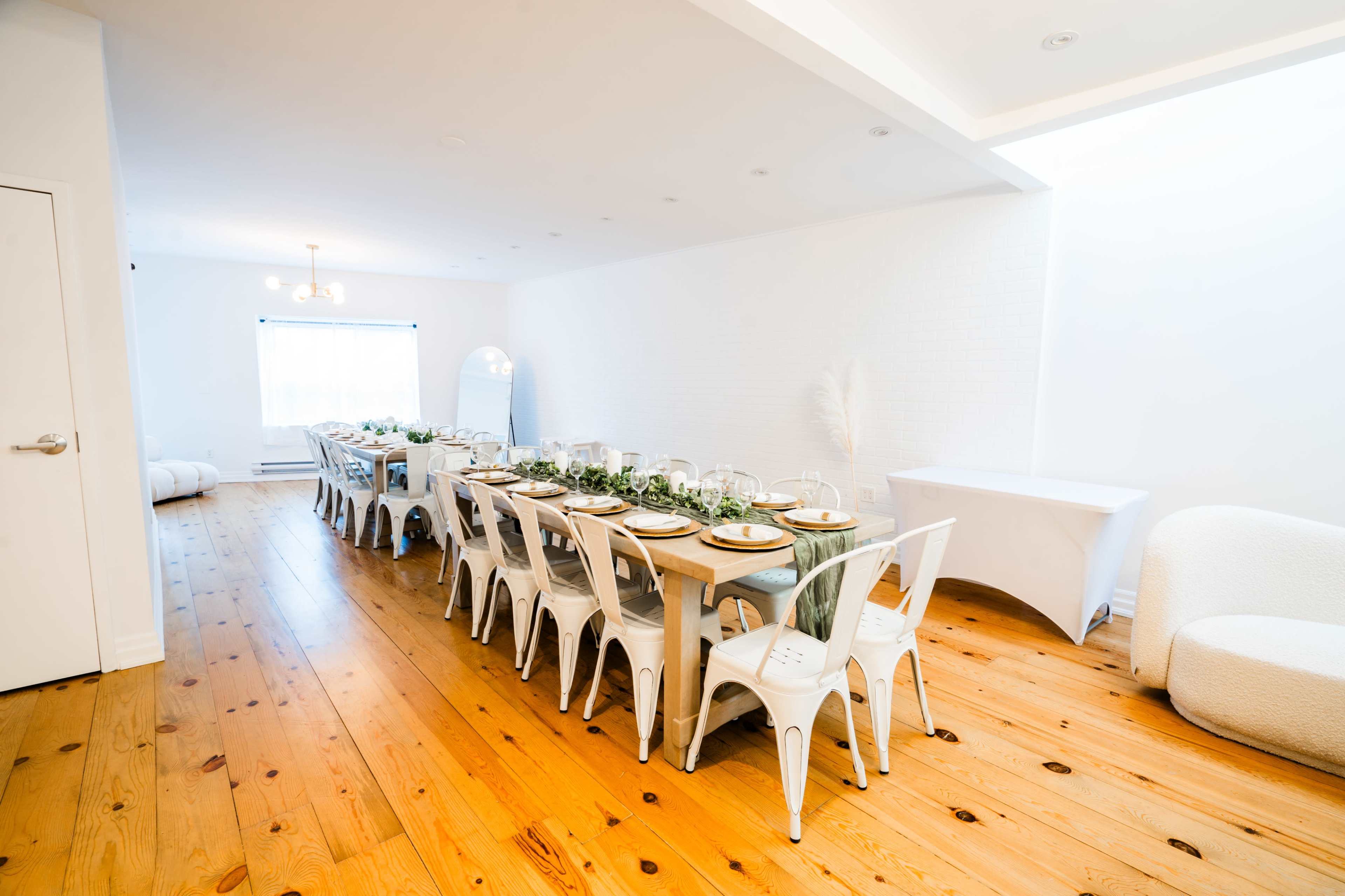 A long dining table with white chairs is set up in a well-lit room featuring wooden floors and minimalistic decor.