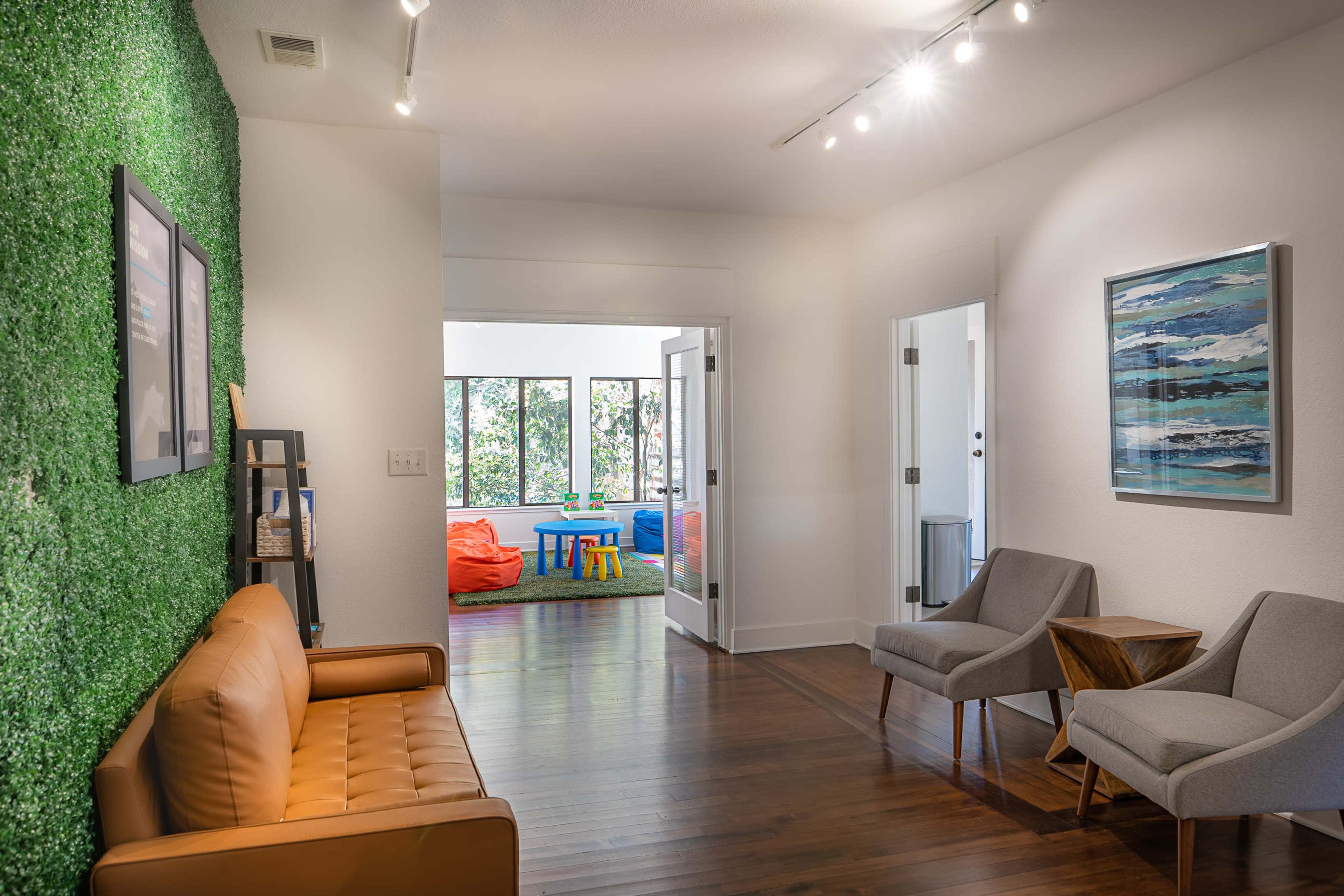 A modern interior space featuring a green wall, a brown leather sofa, two gray chairs, and a doorway leading to a brightly lit room with colorful furniture.
