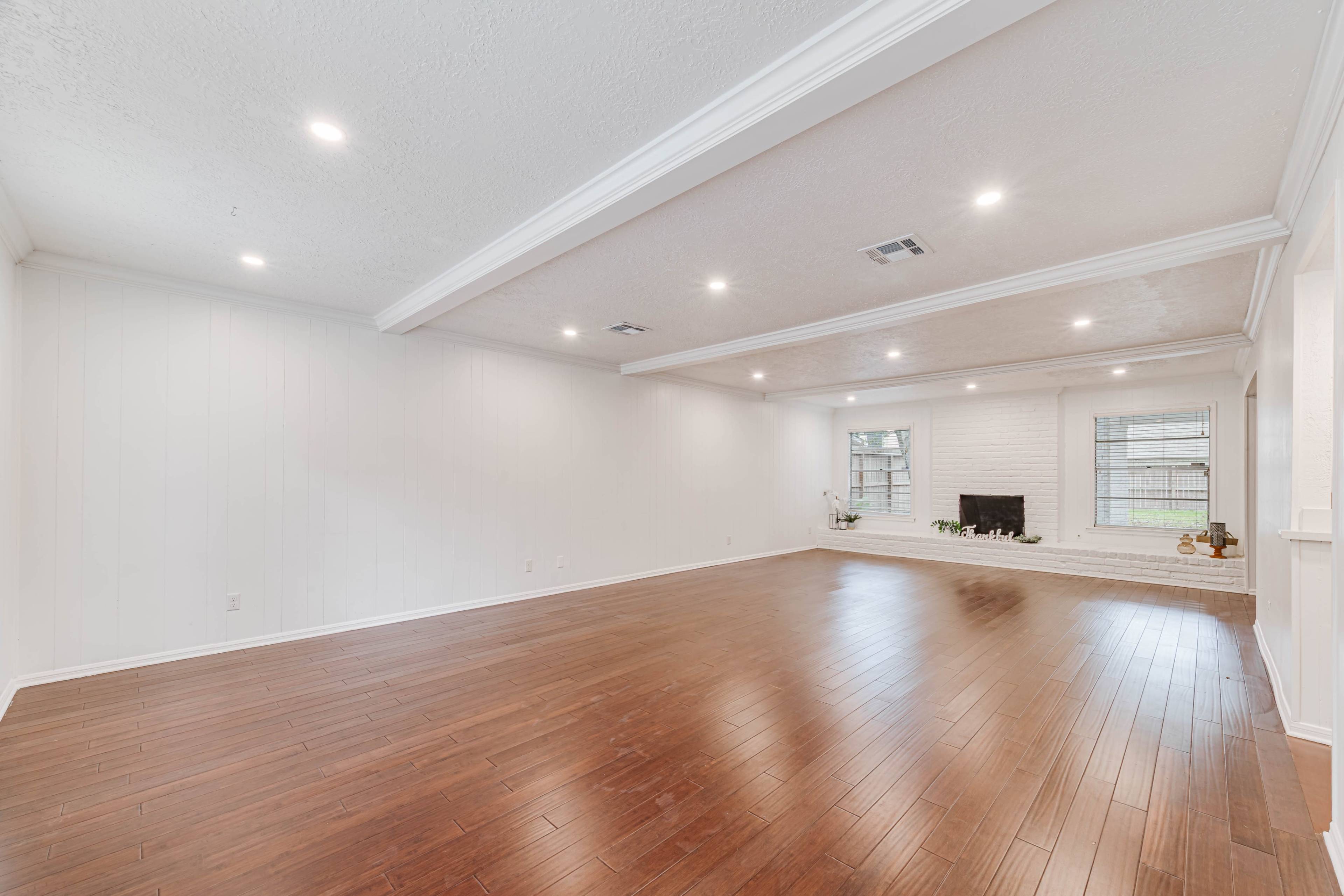 A spacious living room with hardwood flooring, white walls, recessed lighting, and a brick fireplace.