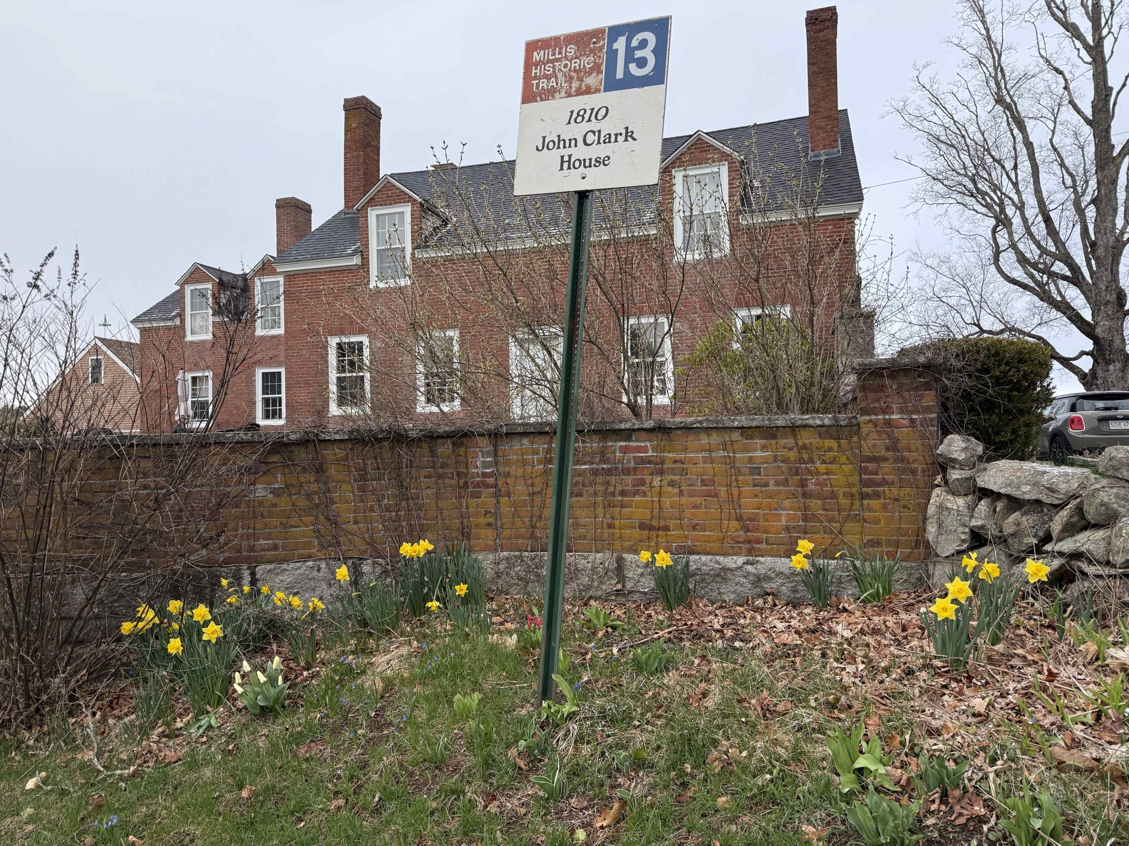 The image shows a historical sign for the John Clark House, marked with the year 1810, alongside a garden of blooming daffodils in front of a large brick building.