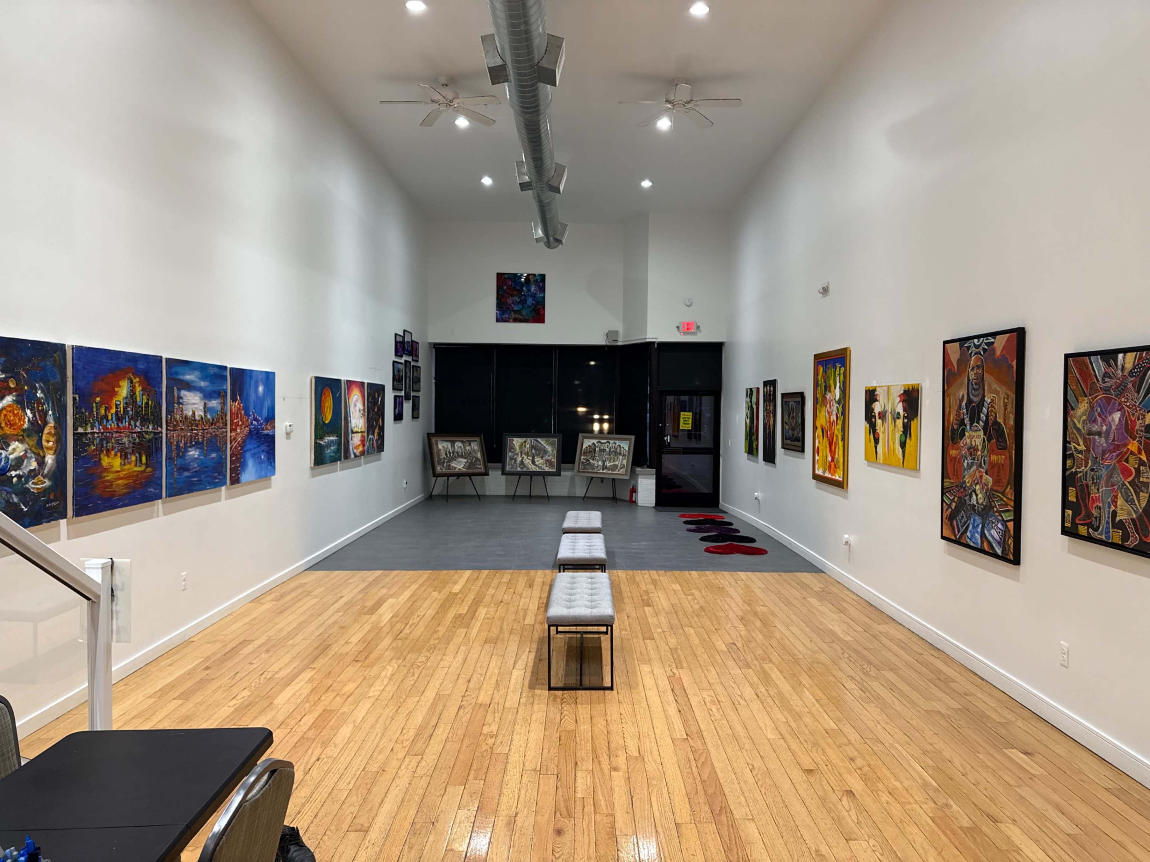The image depicts an art gallery with various colorful paintings displayed on the walls, illuminated by bright overhead lights, and featuring a wooden floor and benches in the center.